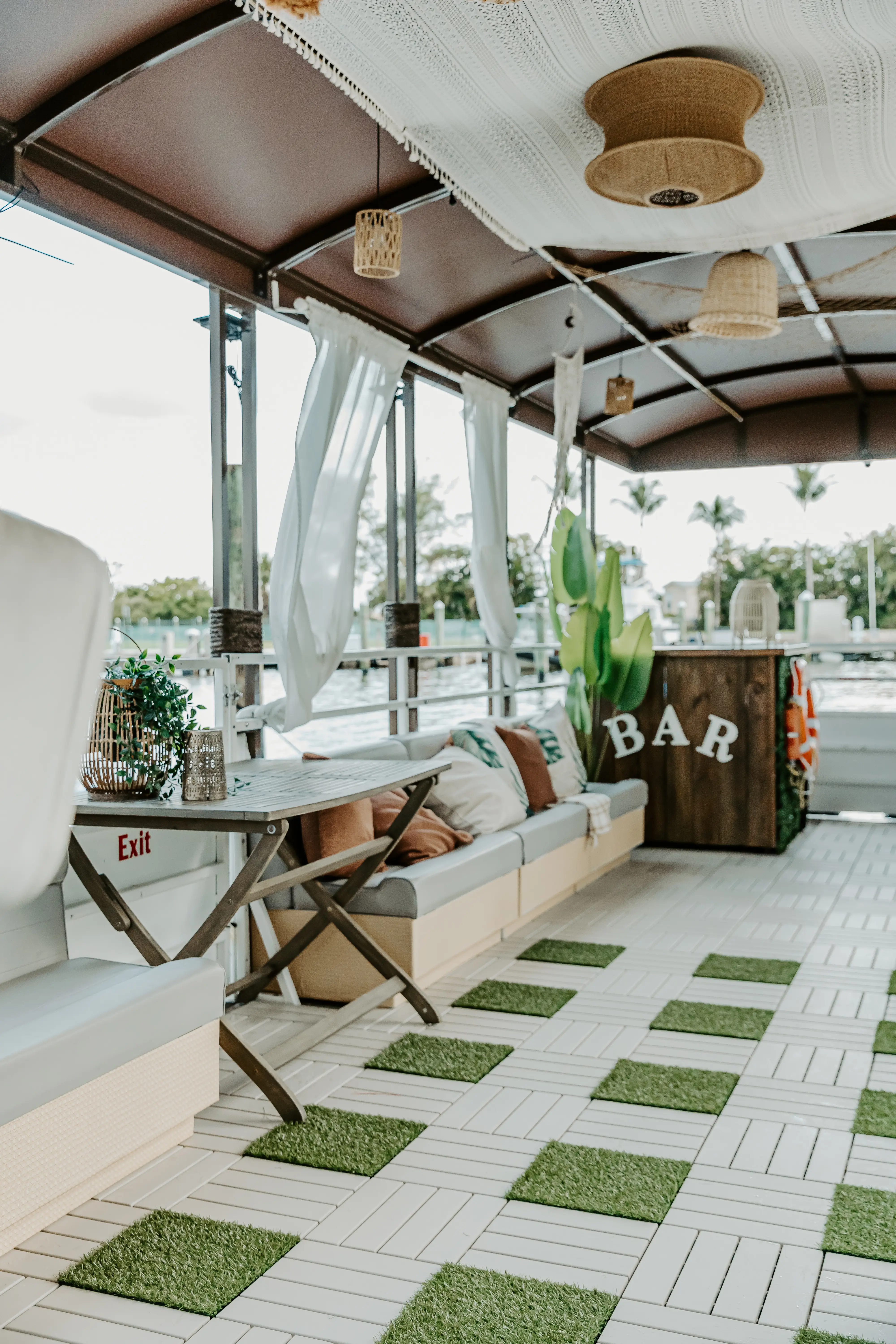 Close-up of the Elite Sport Yachts 45ft Pontoon interior showing the wooden mini-bar station labelled BAR, comfortable gray cushioned bench seating with decorative pillows, wicker hanging lanterns, and the checkered white tile and artificial grass deck floor
