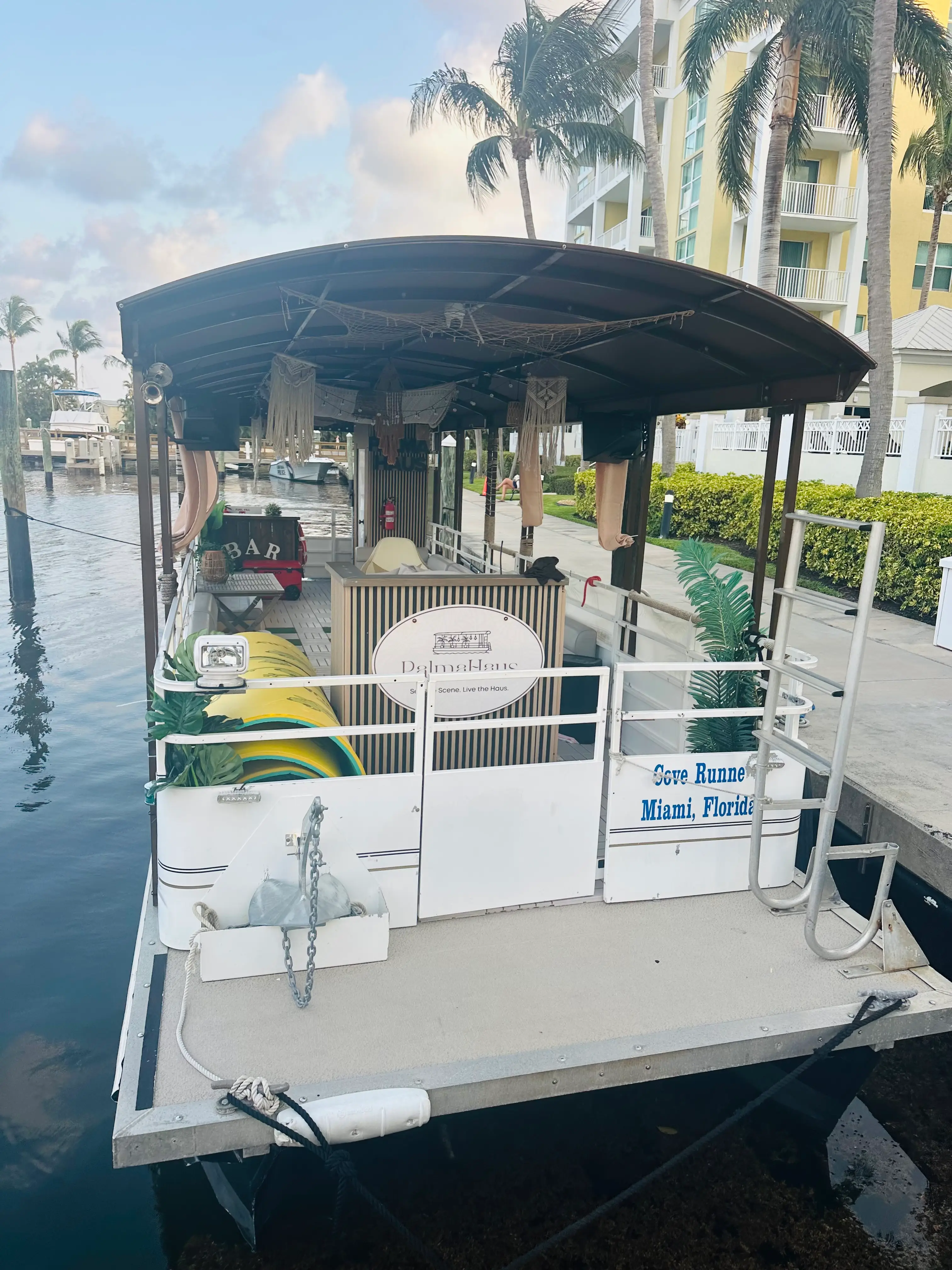 Stern boarding area of the Elite Sport Yachts 45ft Pontoon docked at a canal marina, showing colorful water toy floats stowed on deck, macramé ceiling décor, and the vessel registration reading 'Cove Runner, Miami, Florida'