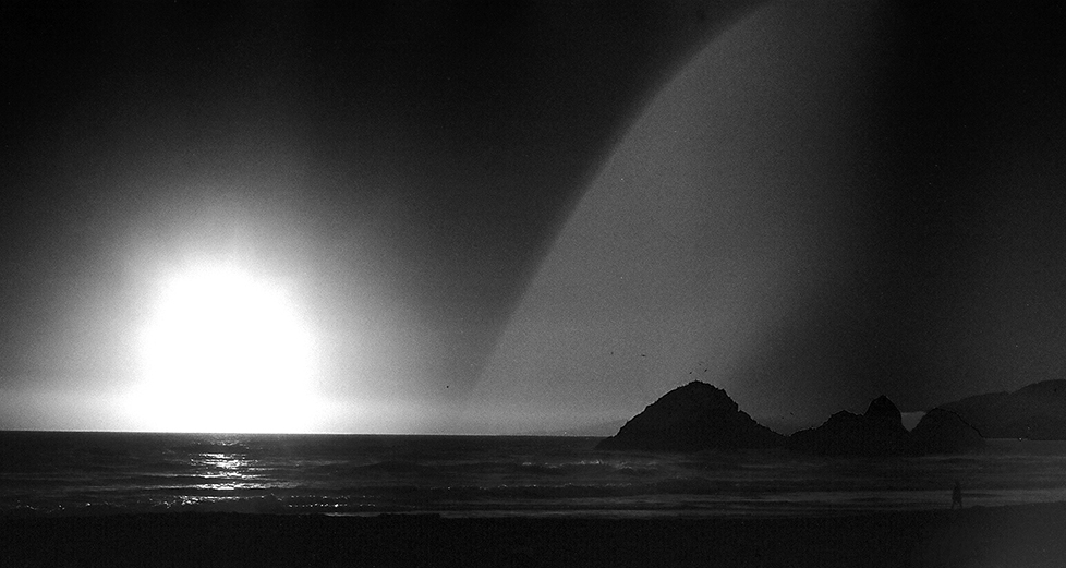 Moody Seal Rocks coastline silhouetted against bright sunset over ocean with rainbow arc in atmospheric sky