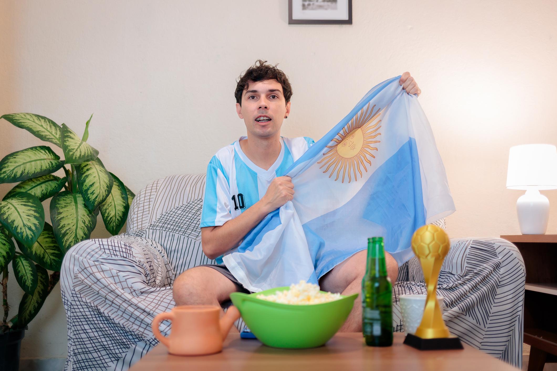 Hincha argentino con camiseta y bandera viendo partido en casa con pochoclo y cerveza