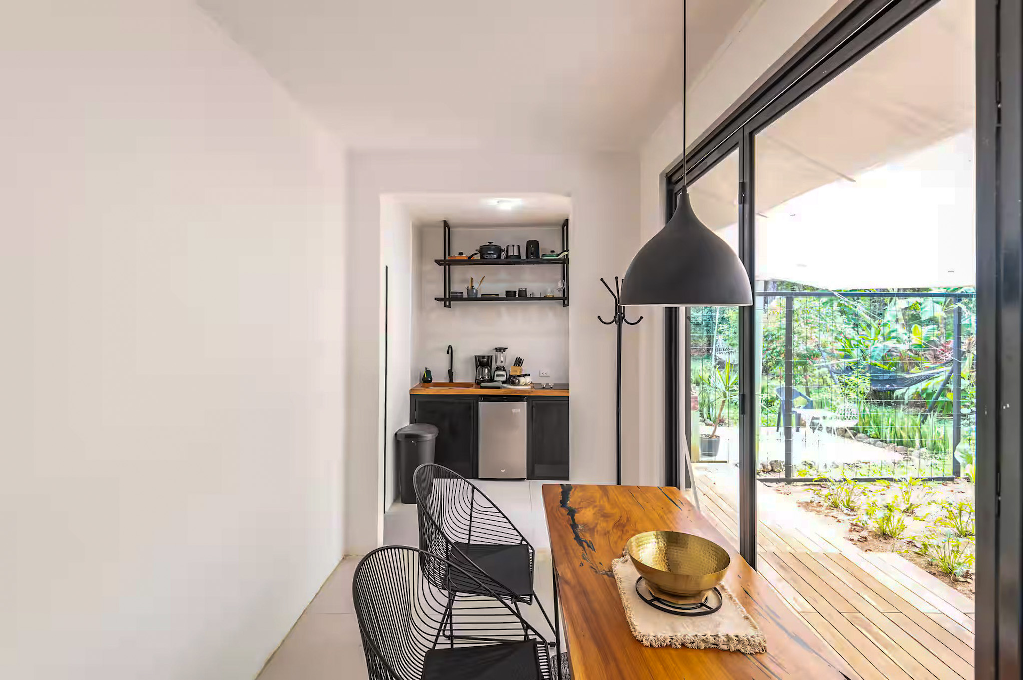 Modern kitchen and dining area with black pendant lamp, wooden table, metal chairs, open shelving, and garden views