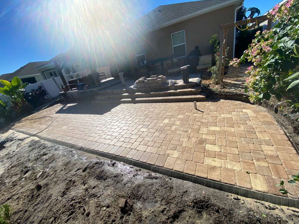 Tan and beige cobblestone paver patio with raised steps leading to an outdoor seating area under bright sunlight