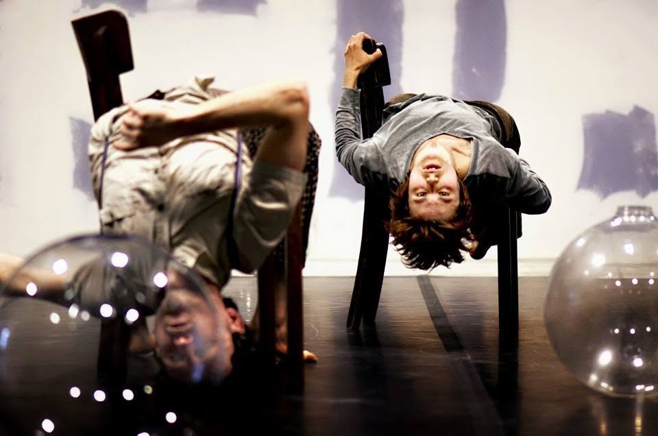 Two performers balancing upside down on chairs with glass spheres