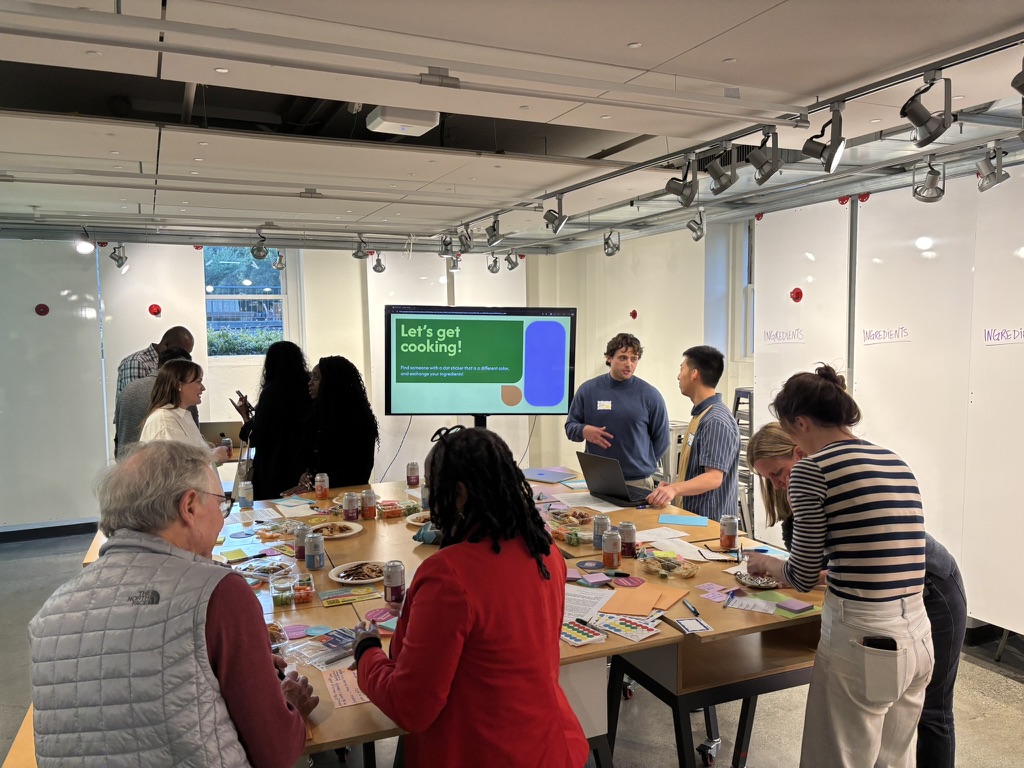 Monitor displaying "Let's get cooking!" slide, participants eating around tables