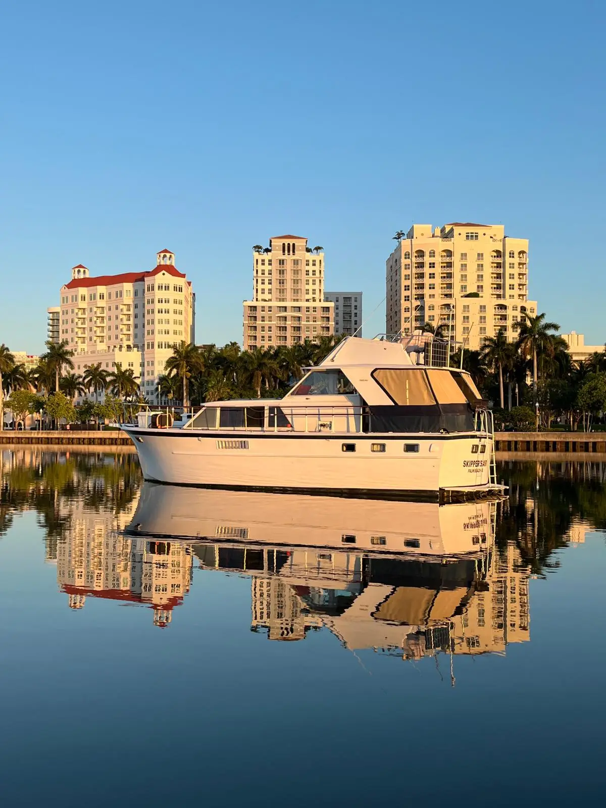 The Elite Sport Yachts 50ft Hatteras Motor Yacht 'Skipper's Joy' reflected perfectly in glassy West Palm Beach waterway at golden hour, with the downtown skyline and palm trees in the background