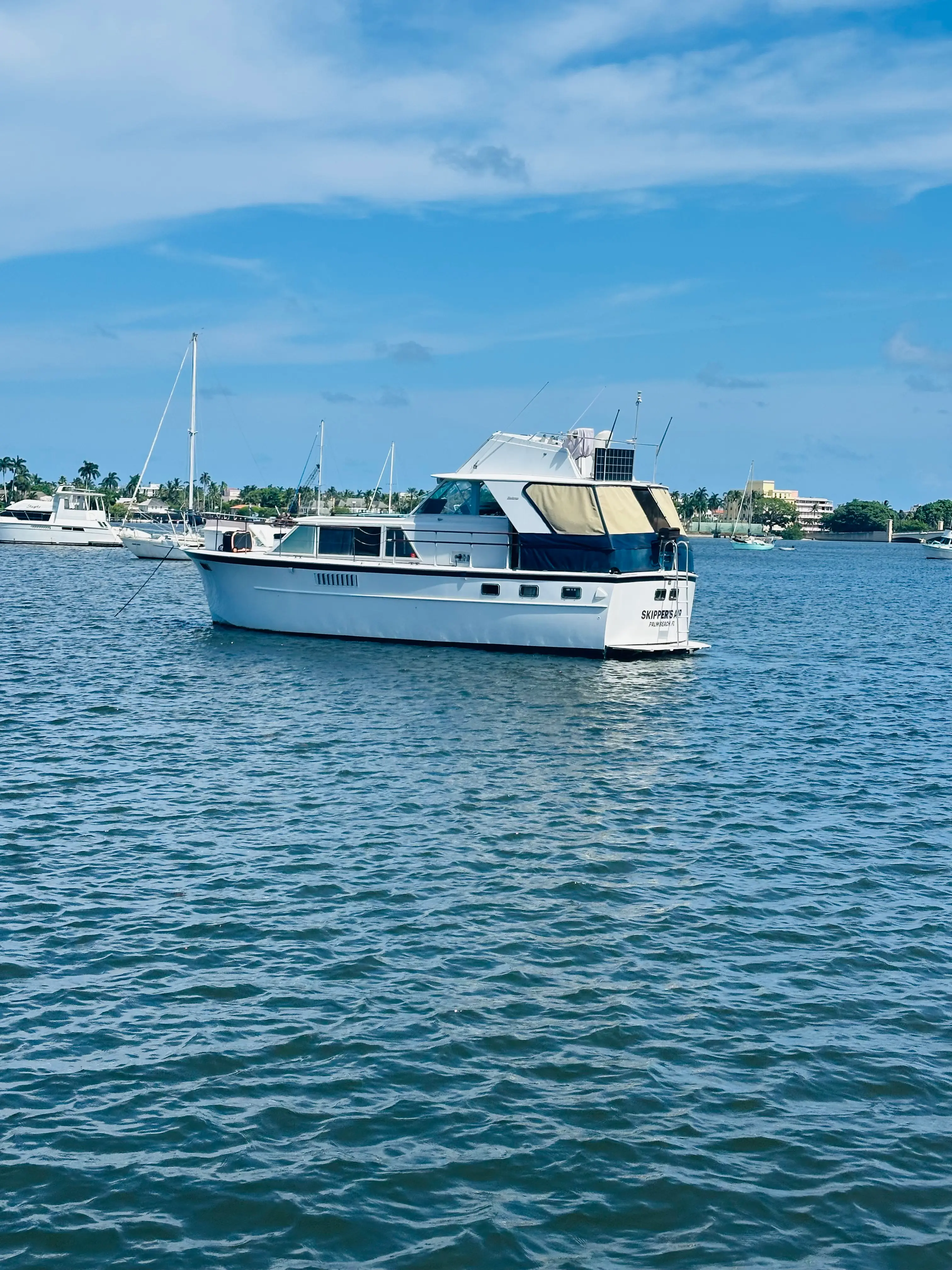 Full side profile of the Elite Sport Yachts 50ft Hatteras Motor Yacht anchored in open West Palm Beach waters on a sunny day, with sailboats and marina visible in the distance