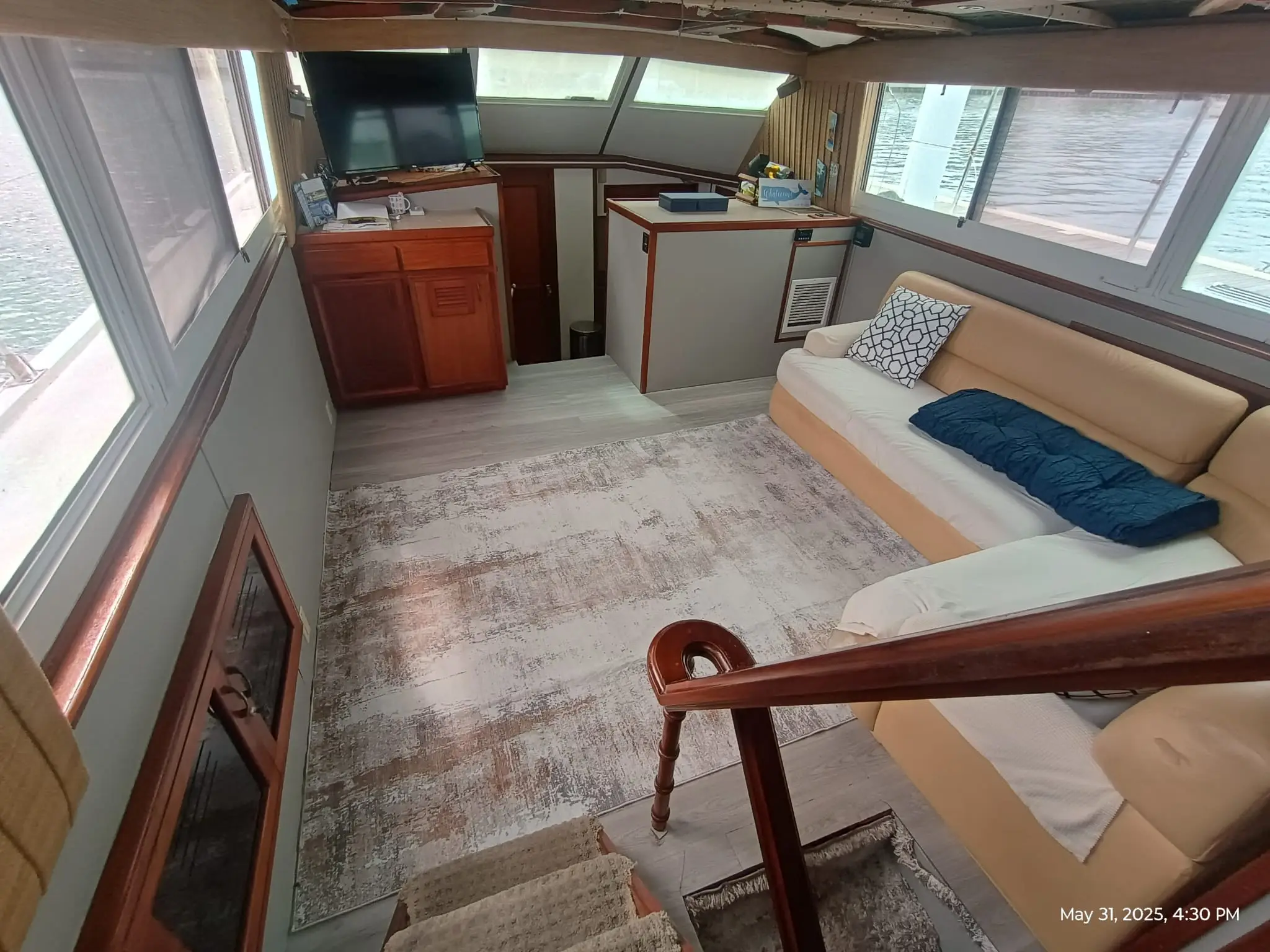Interior lounge and galley area of the Elite Sport Yachts 50ft Hatteras Motor Yacht showing cream upholstered sofa seating, a mounted flatscreen TV, warm wood cabinetry, built-in refrigerator, and large panoramic windows overlooking the water