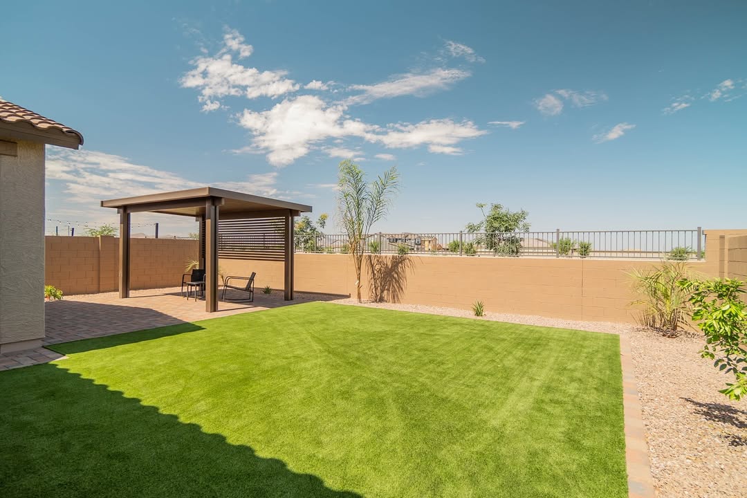 Wide backyard view of bronze pergola and brick paver patio with large artificial turf lawn, desert shrubs, and block wall fence