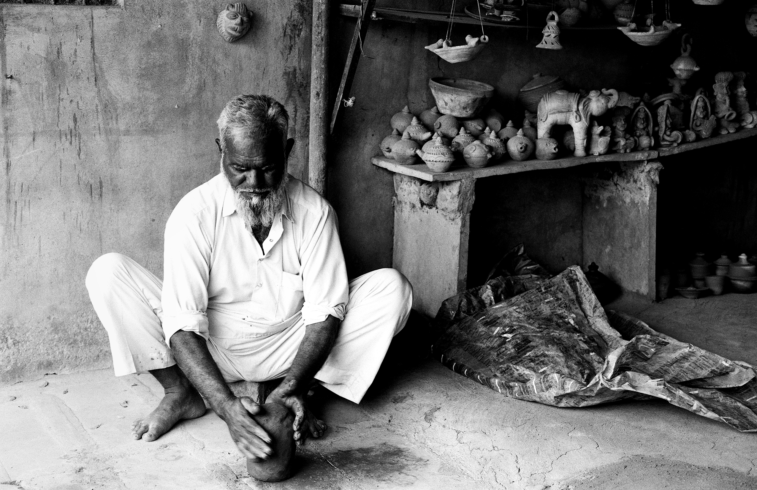 Elderly bearded man in white clothing sitting by clay pottery shop