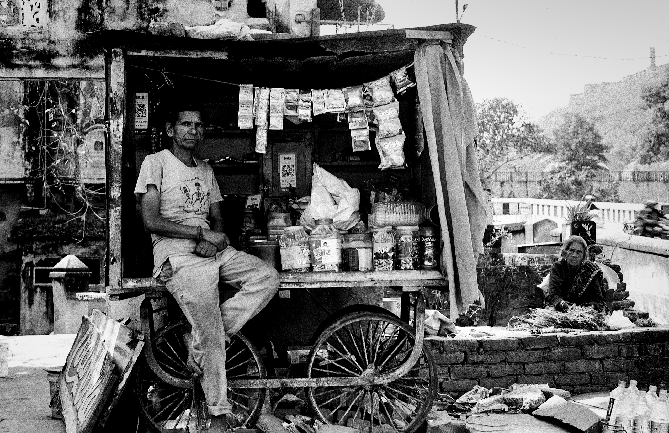 Street vendor at chai cart with woman sitting nearby in urban Indian street