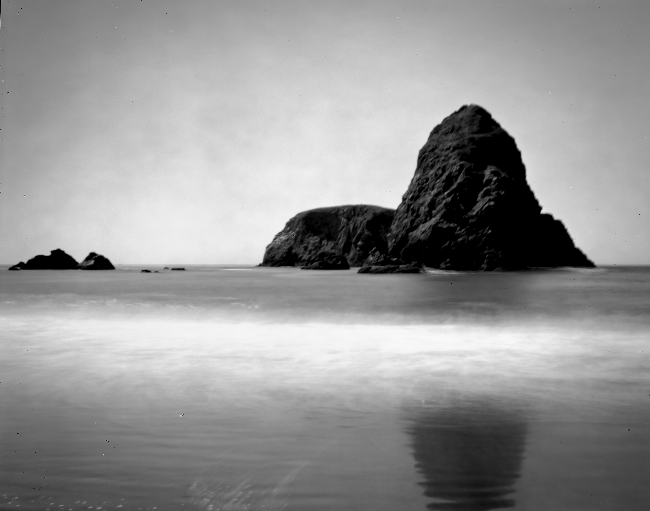Dramatic sea stack rock formation rising from ethereal misty long-exposure water with soft reflection under overcast coastal sky