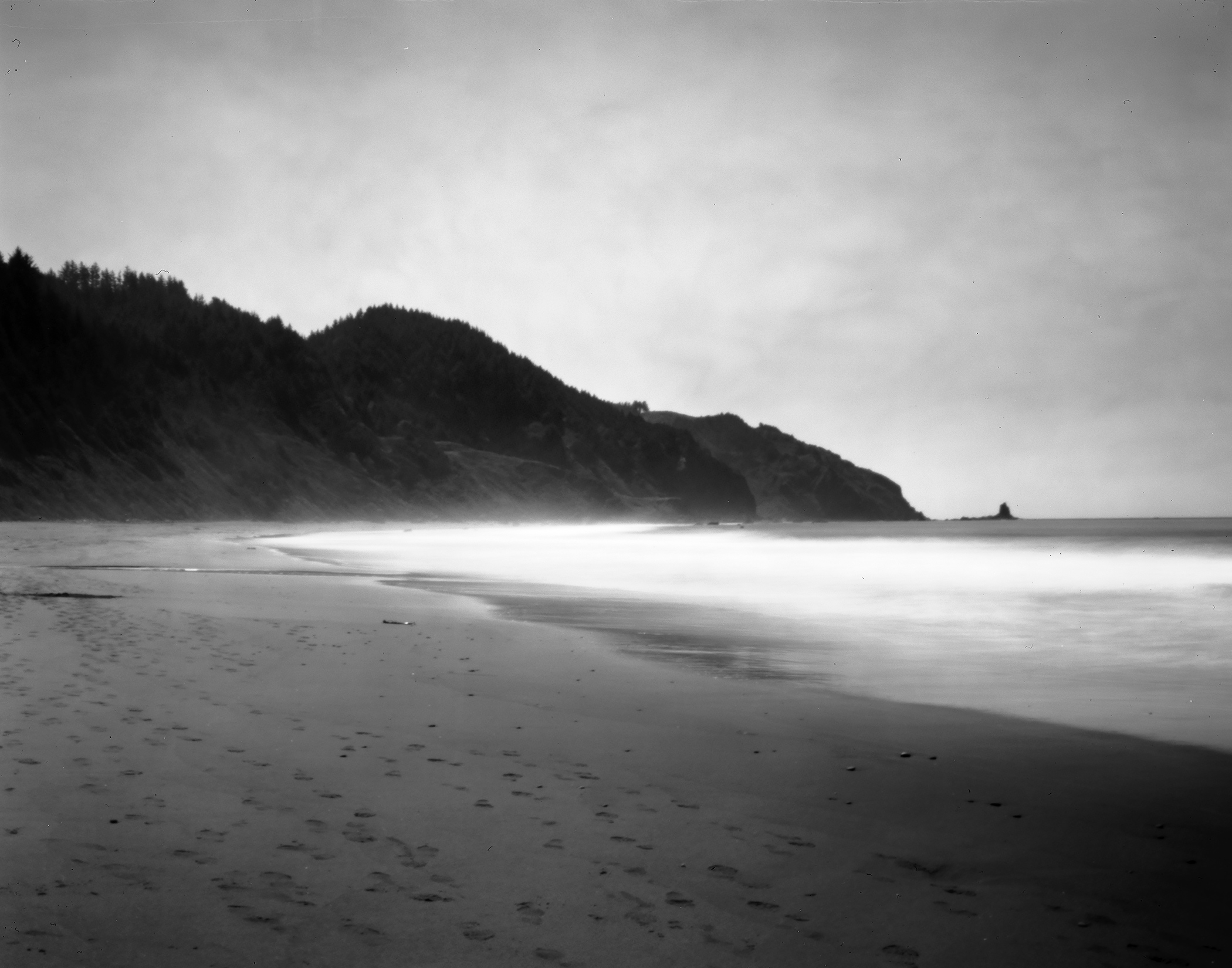 Moody coastal beach with footprints in sand curved shoreline forested mountains and dreamy long-exposure misty water
