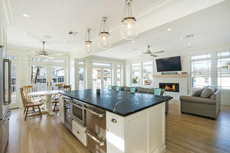 Large white kitchen island with dark countertop over light oak hardwood and fireplace beyond