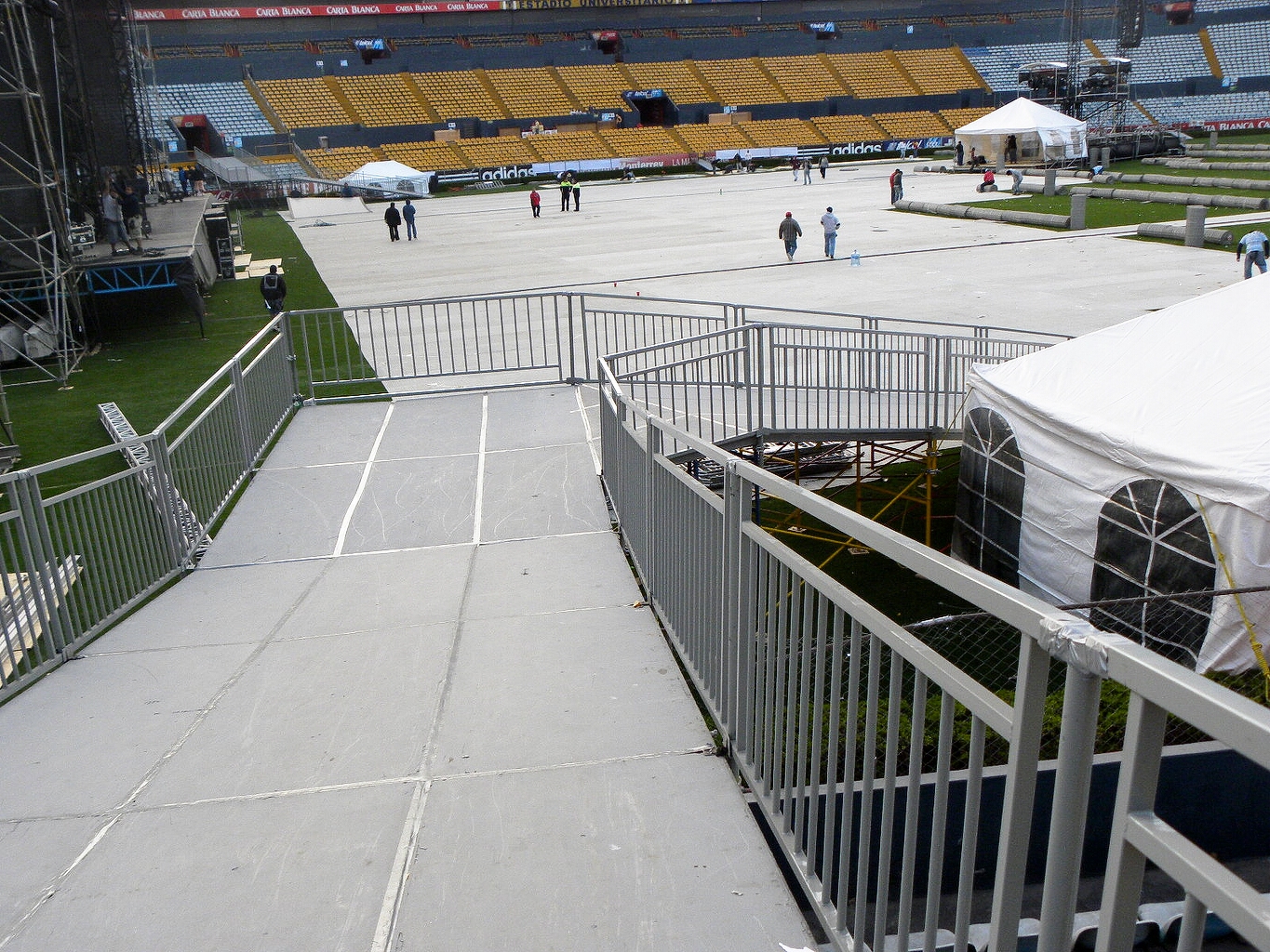 Múltiples tarimas blancas en estadio profesional con gradas amarillas, carpas blancas, seguridad con barreras metálicas y vista amplia de instalaciones