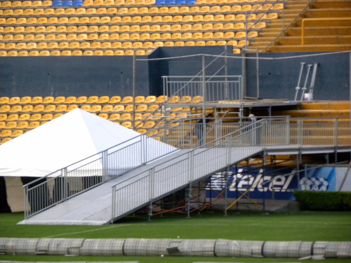Tarima blanca con rampa de acceso profesional en estadio con gradas amarillas y estructura de barandales metálicos