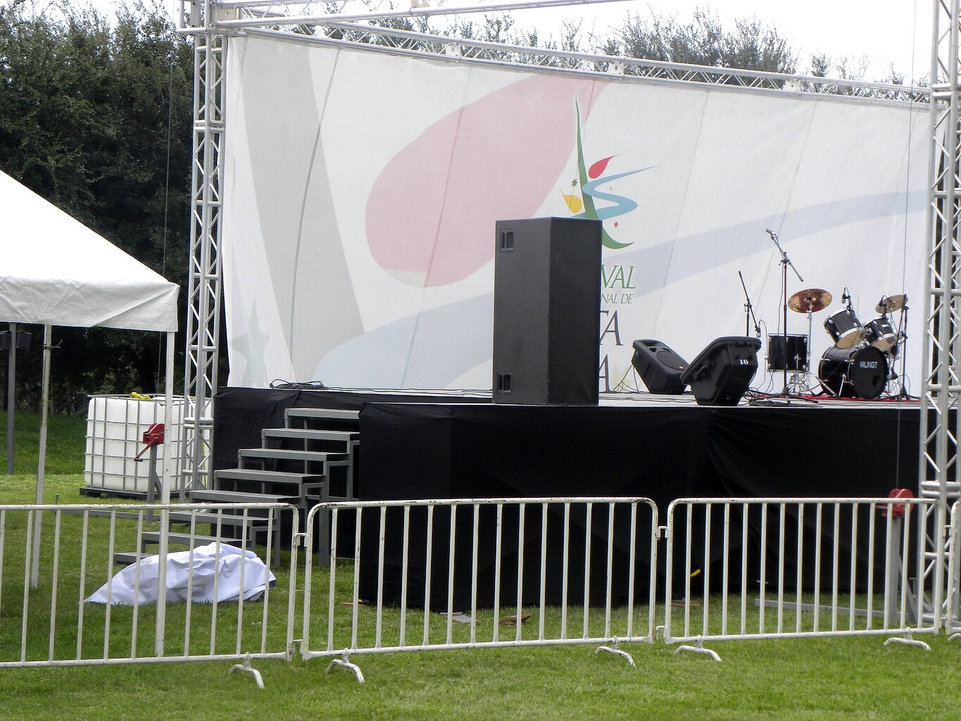 Escenario exterior con estructura de truss metálica, carpa blanca, panel decorativo colorido, plataforma negra elevada y equipo de sonido profesional para festival