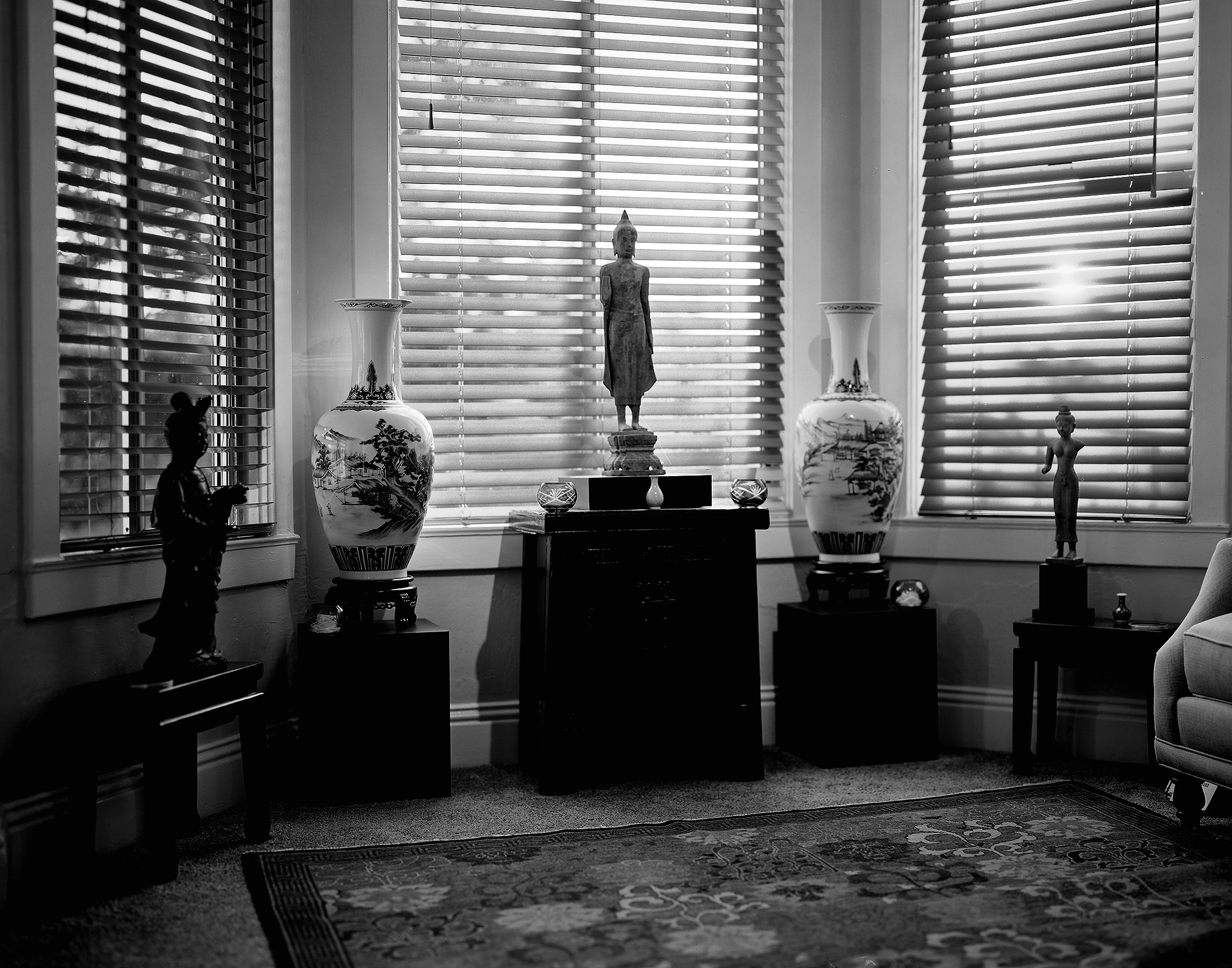 Elegant interior composition with Buddhist and Asian statues arranged on black pedestals against large windows with venetian blinds casting geometric shadows