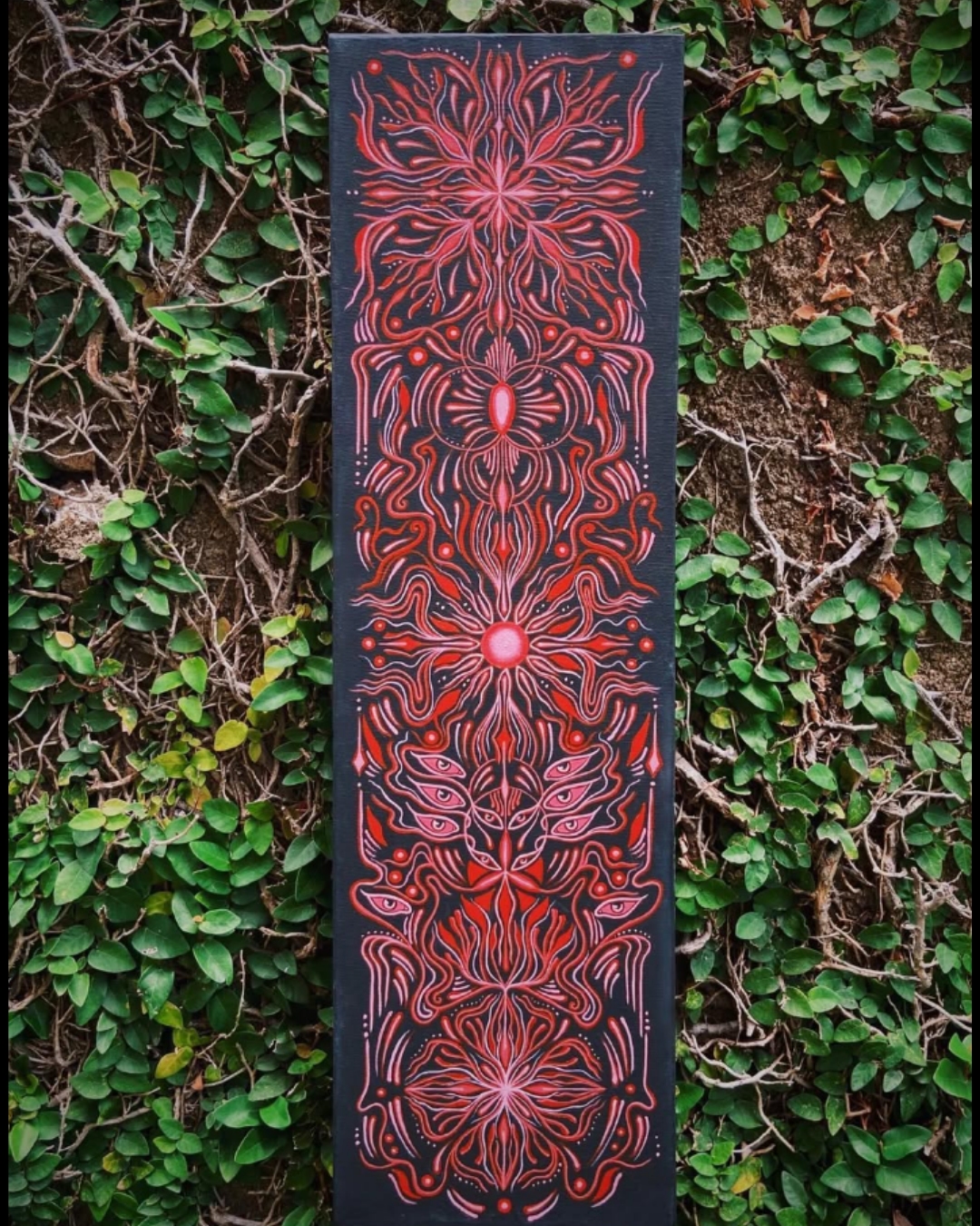 Pintura vertical preta com grafismos vermelhos simétricos e olhos sobre folhagem