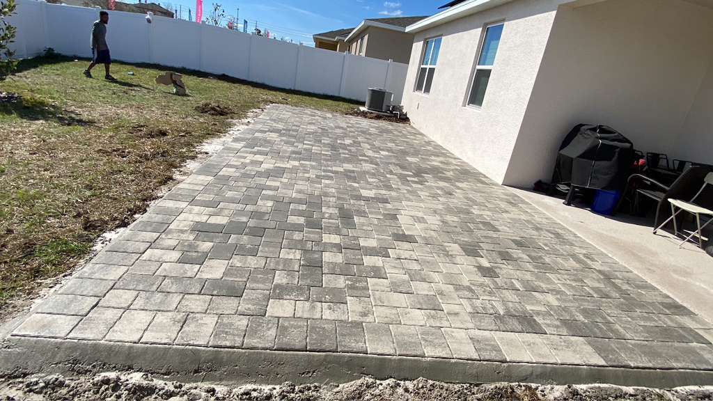 Grey herringbone brick paver pathway along the side of a white stucco home with white vinyl fence