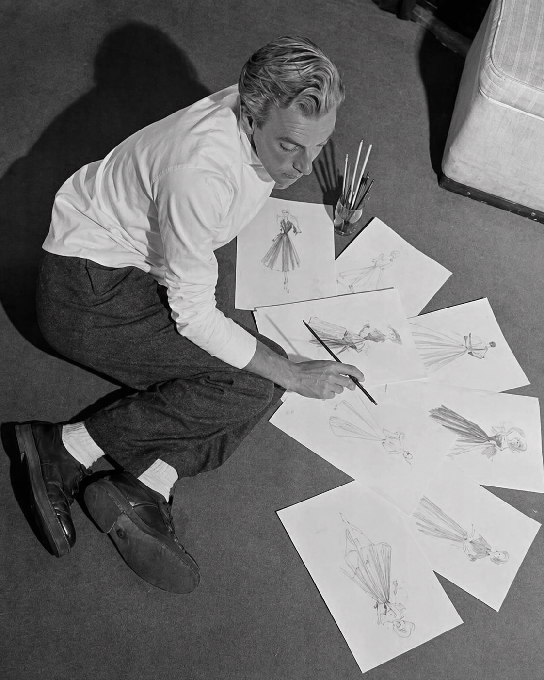 Jacques Fath seated on the floor surrounded by fashion sketches, drawing with pencil, black and white