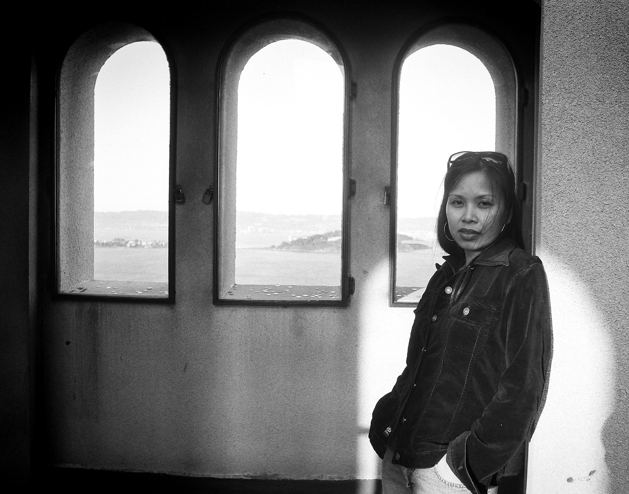 Woman in denim jacket standing by arched windows overlooking San Francisco Bay with natural light