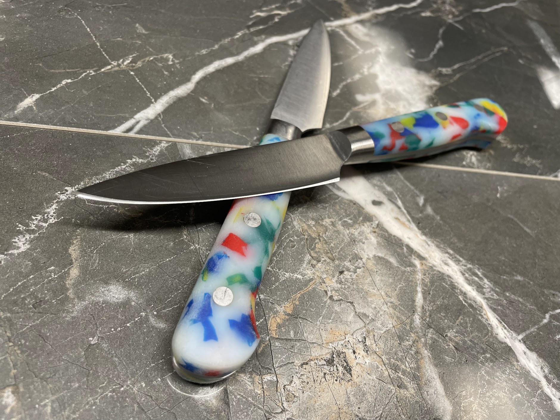 Two paring knives with colorful speckled resin handles crossed on grey marble