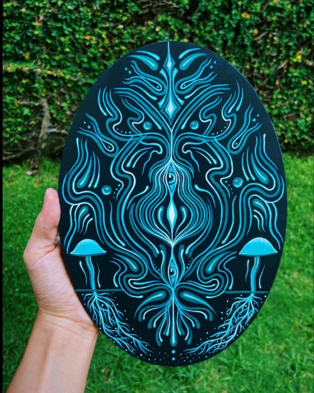 Pintura oval preta com grafismos em turquesa e cogumelos azuis segurada por mão na grama