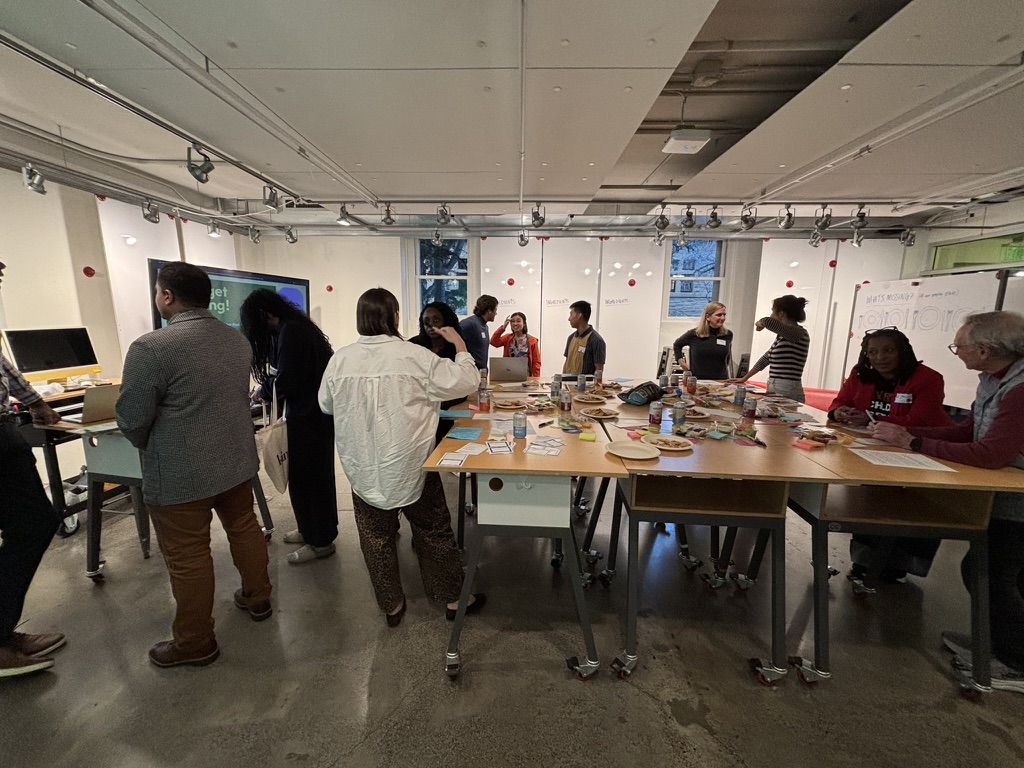 Wide view of d.school event space with approximately 14 participants standing and mingling