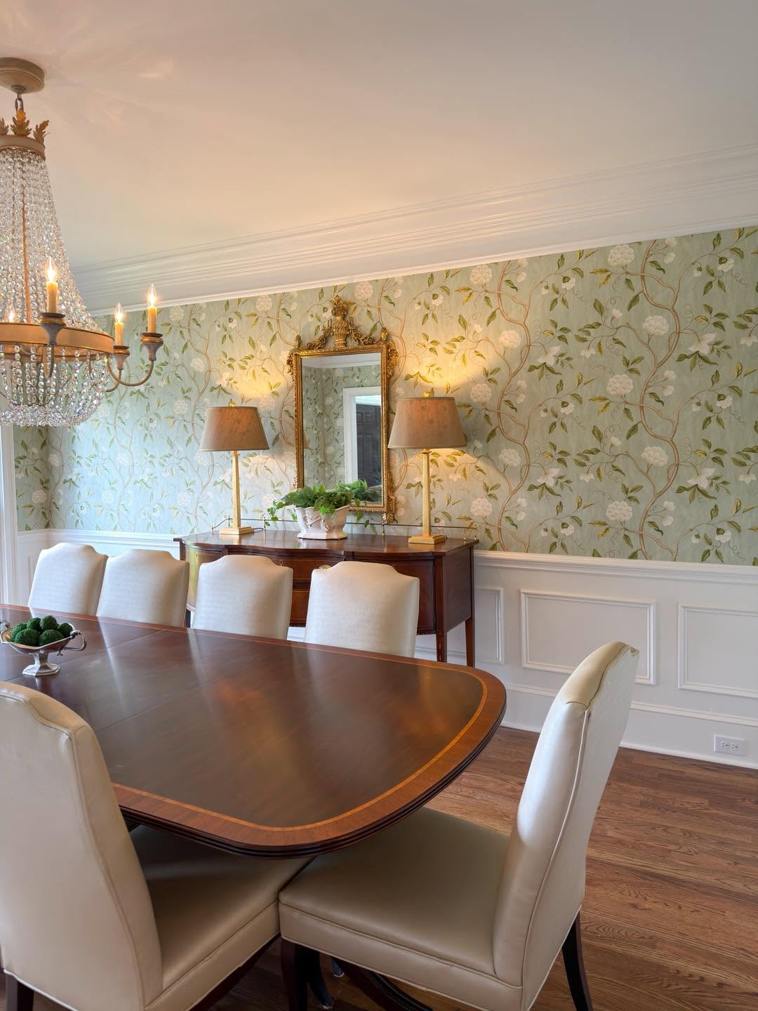 Dining room with hand-installed green floral chinoiserie wallpaper, a crystal chandelier, and white wainscoting