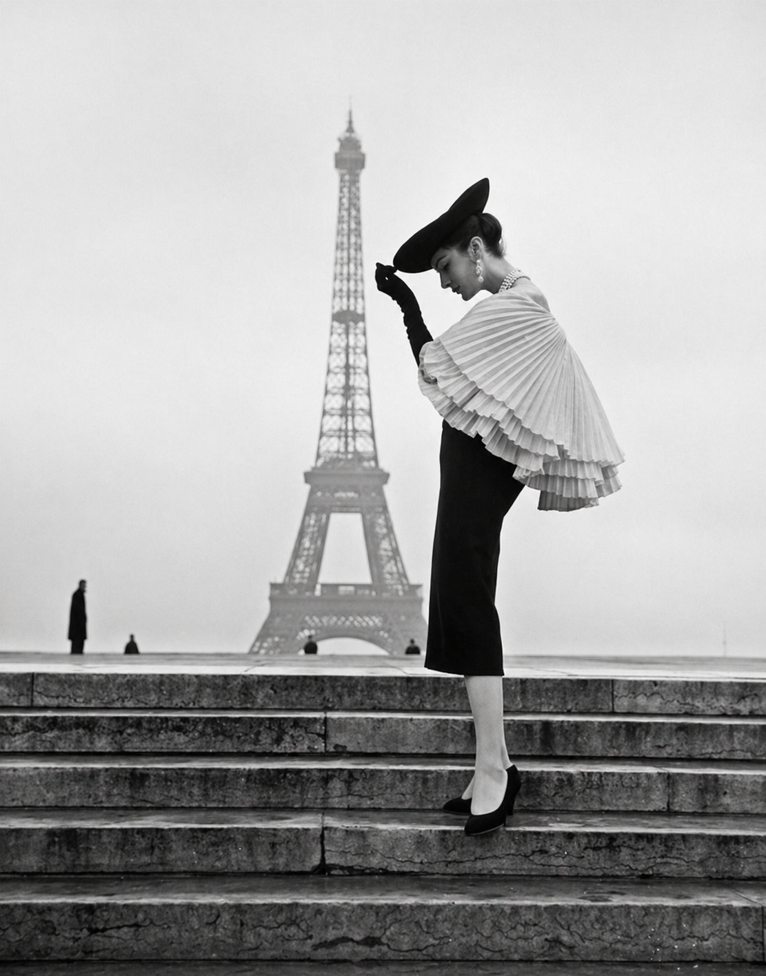 Model in Jacques Fath pleated cape dress on steps before the Eiffel Tower, Paris, black and white