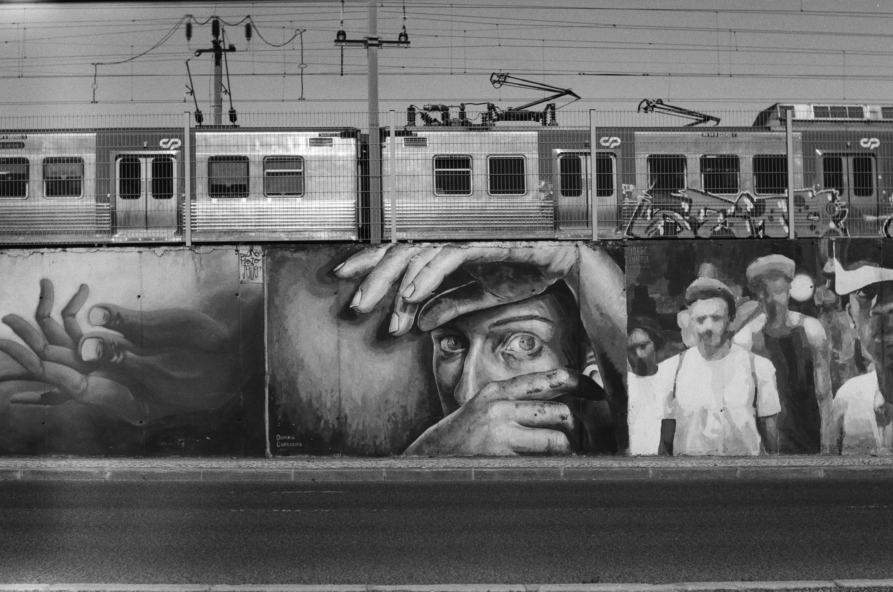 Black and white frame of a commuter train passing above a large street mural of hands and faces