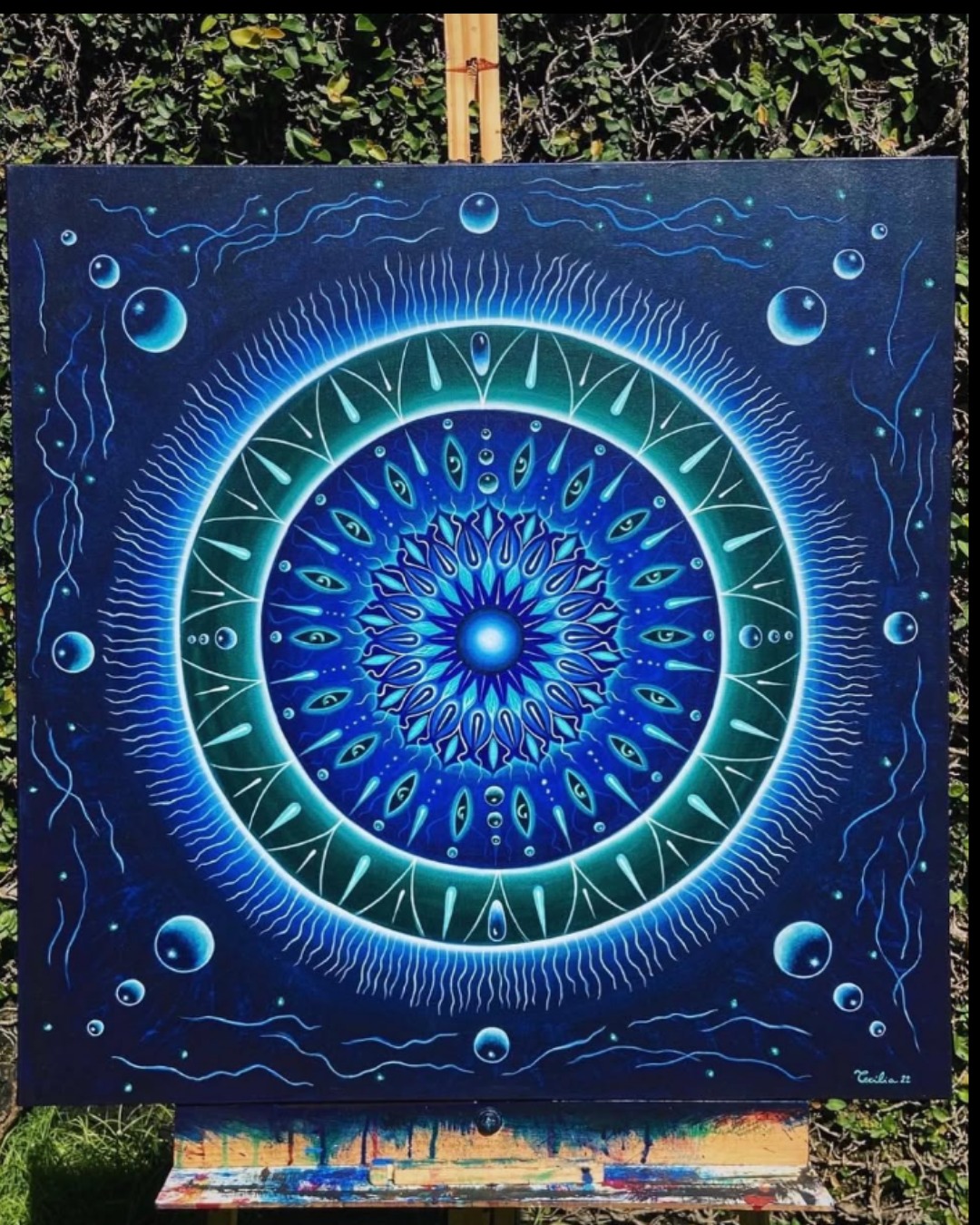 Grande tela quadrada azul escuro com mandala luminosa de olhos e bolhas de água sobre cavalete