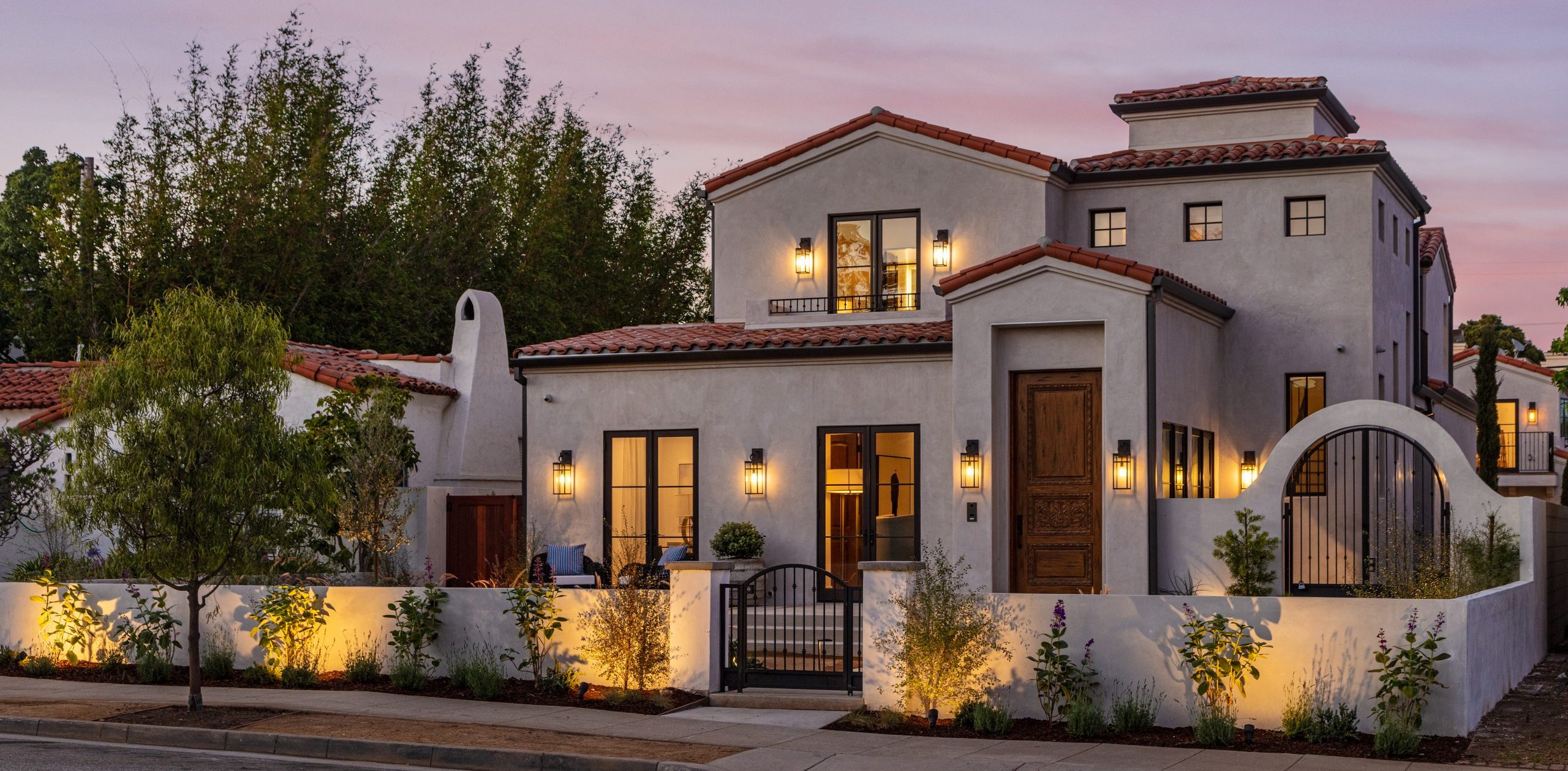 Elegant Mediterranean-style home exterior with warm lighting at twilight, featuring white stucco walls and terracotta roof tiles