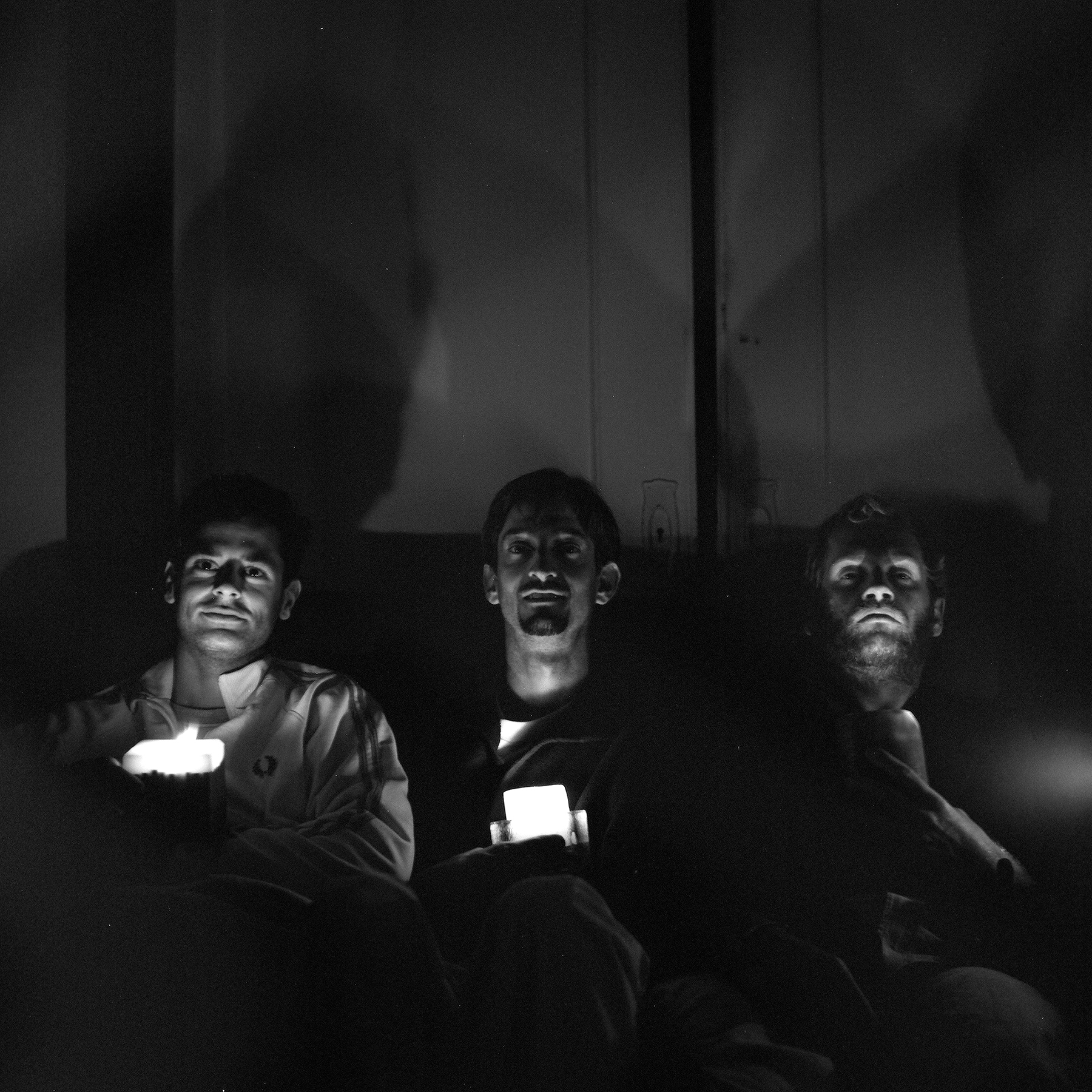 Three men sitting together in darkness faces illuminated by candles creating intimate portrait with dramatic shadows