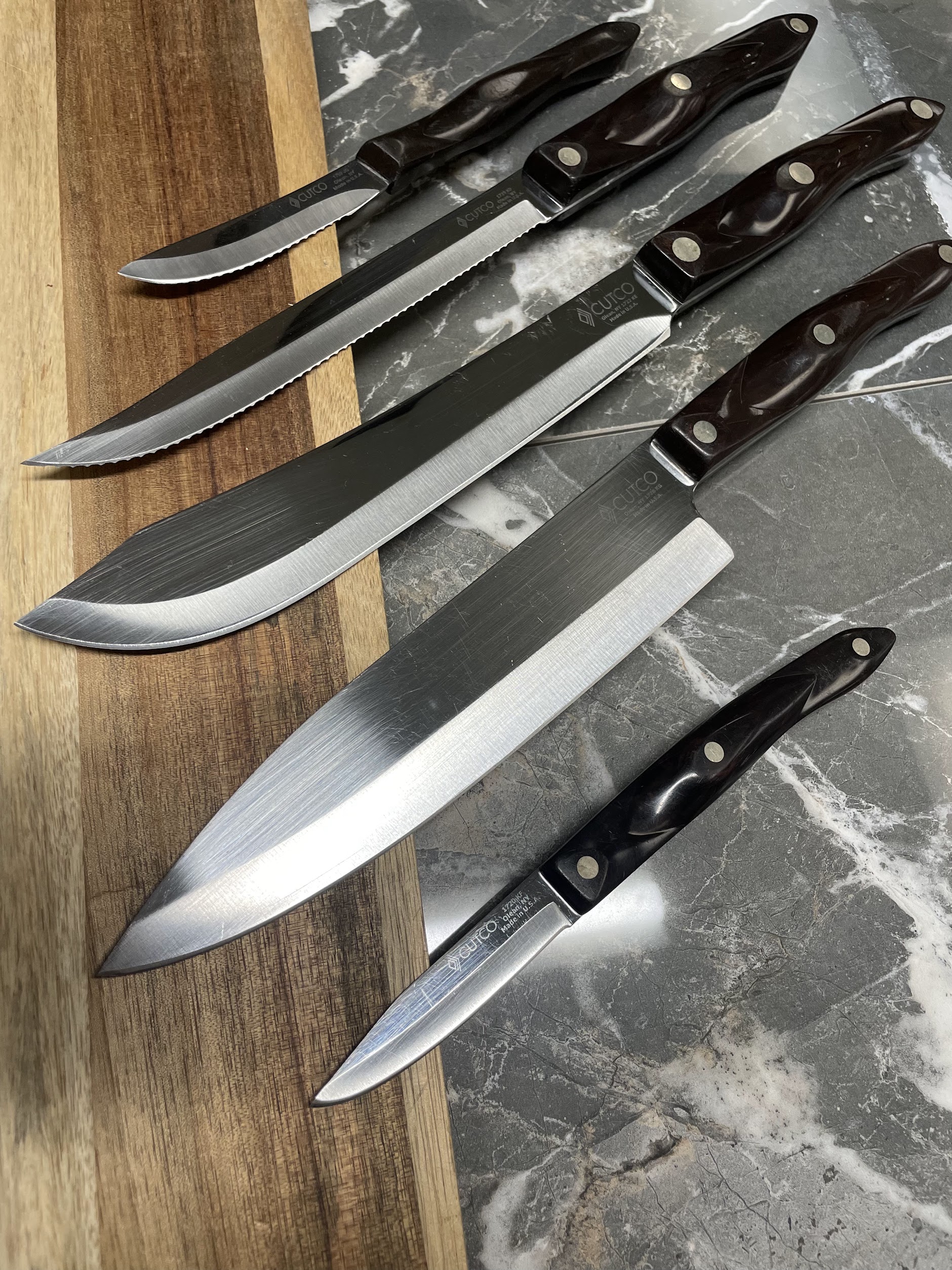 Set of Cutco kitchen knives with dark handles fanned across a walnut board and grey marble