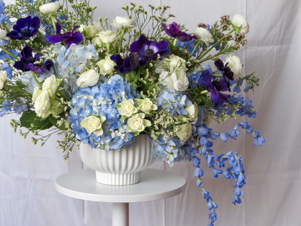 Blue hydrangea, yellow rose, white rose, purple anemone arrangement in white ribbed urn on pedestal