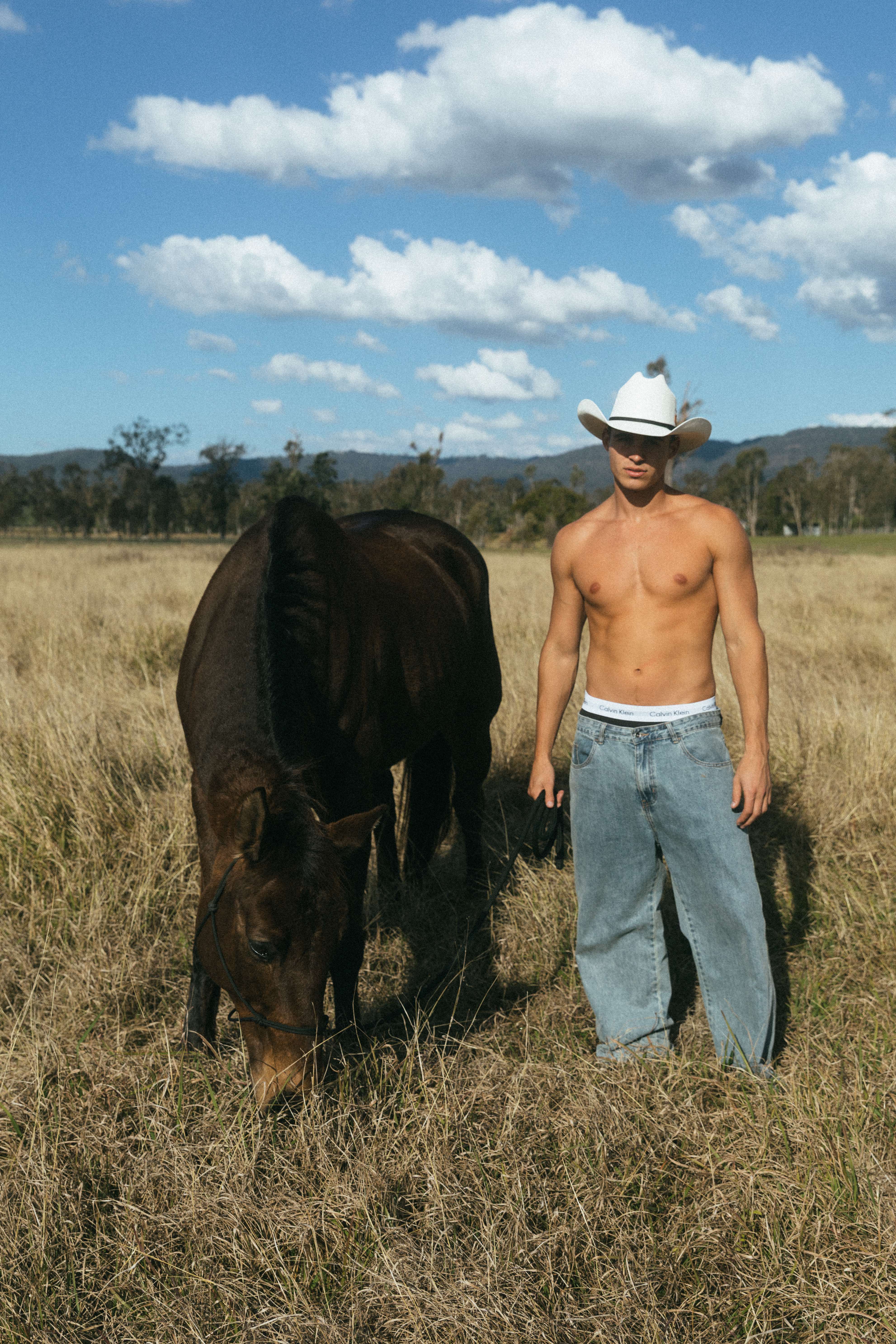 Shirtless cowboy in white hat and jeans standing with bay horse golden field