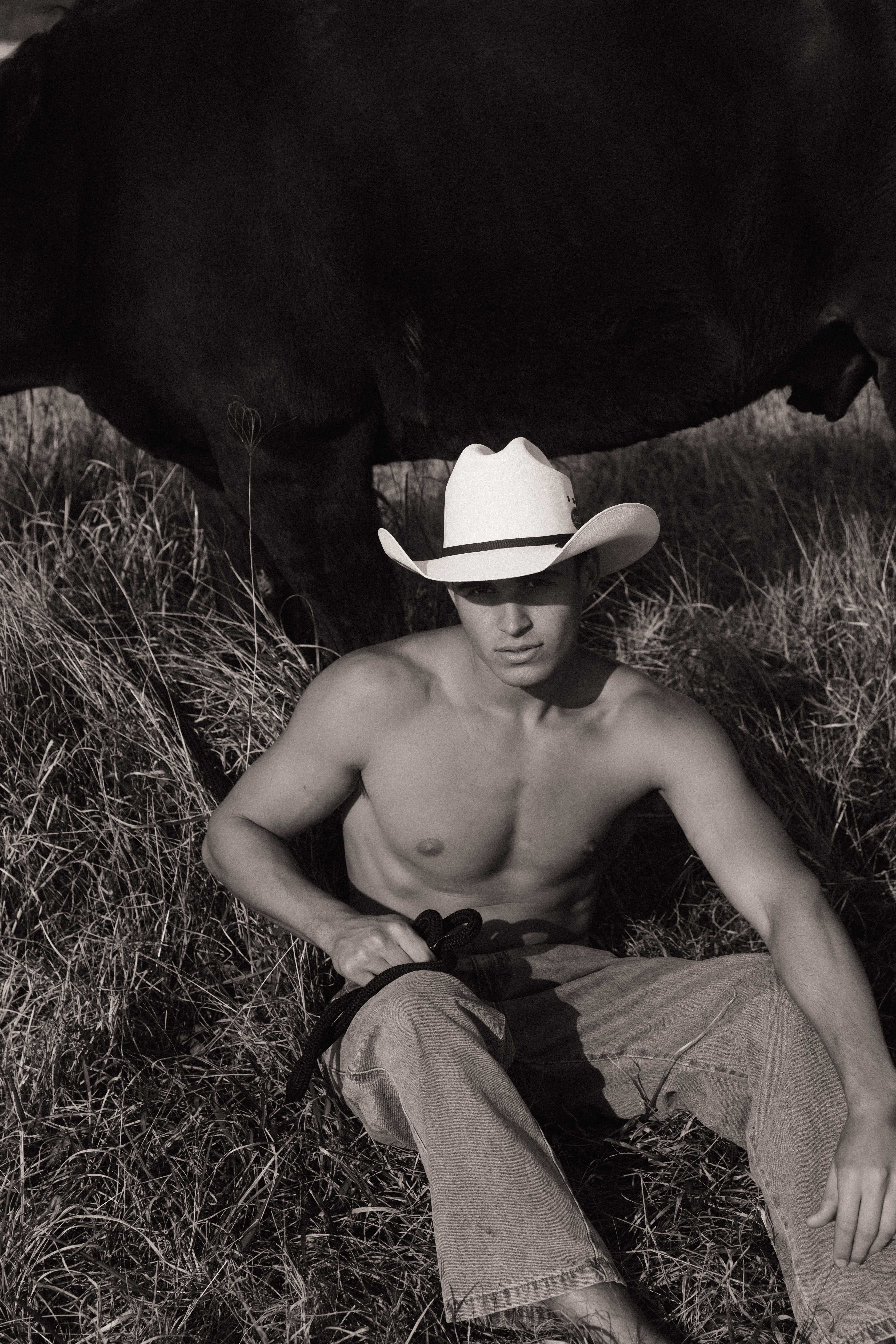 Male model in cowboy hat seated in grass with dark horse black and white