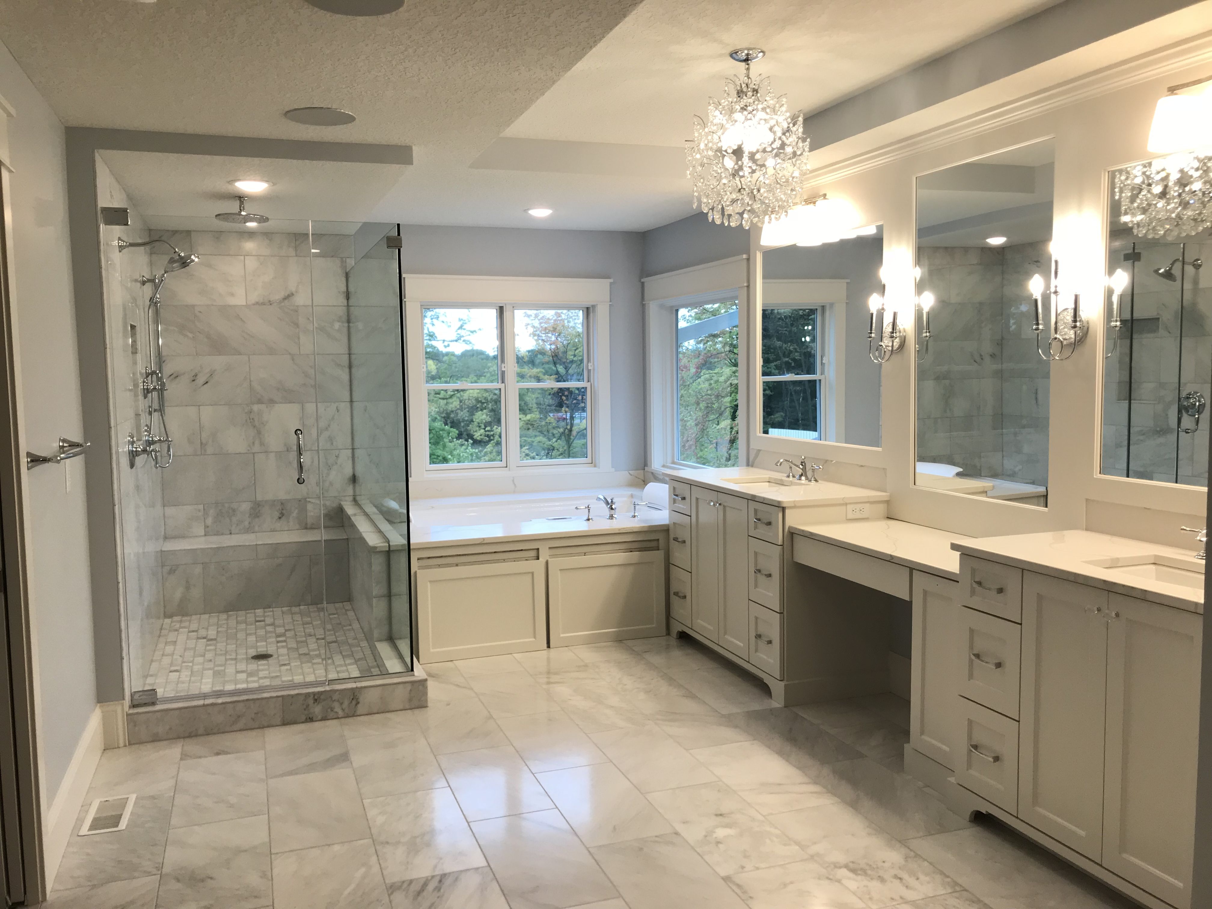 Elegant master bathroom with crystal chandelier, Carrara marble tile, frameless glass shower with bench, and double vanity with chrome fixtures