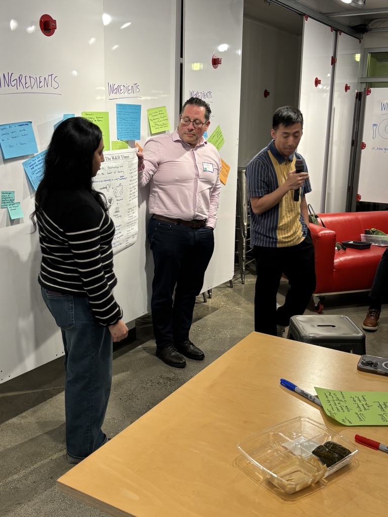 Devin in pink shirt presenting to three participants near whiteboard with INGREDIENTS sticky notes