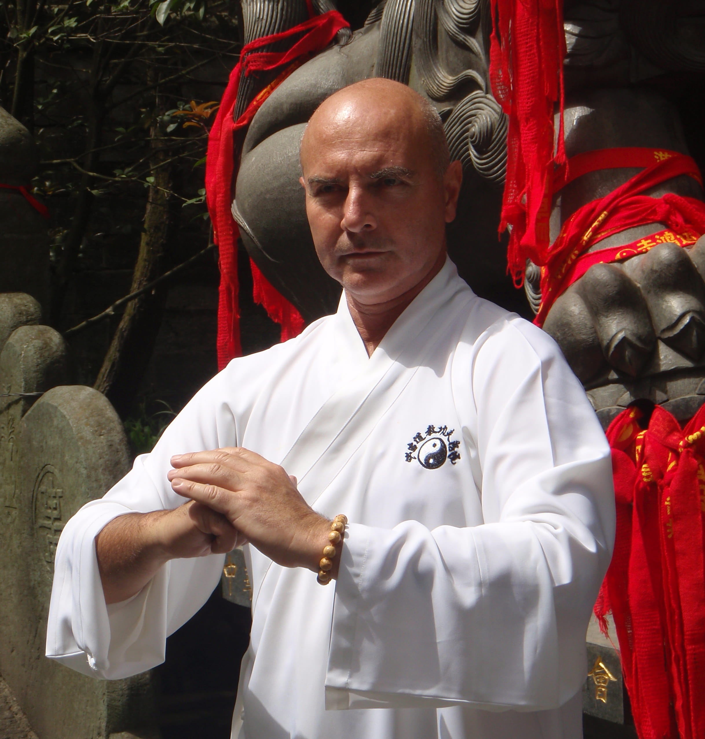 Portrait of Kadama in a white qigong robe, hands held in a salute, in front of a red-ribboned stone guardian statue in China