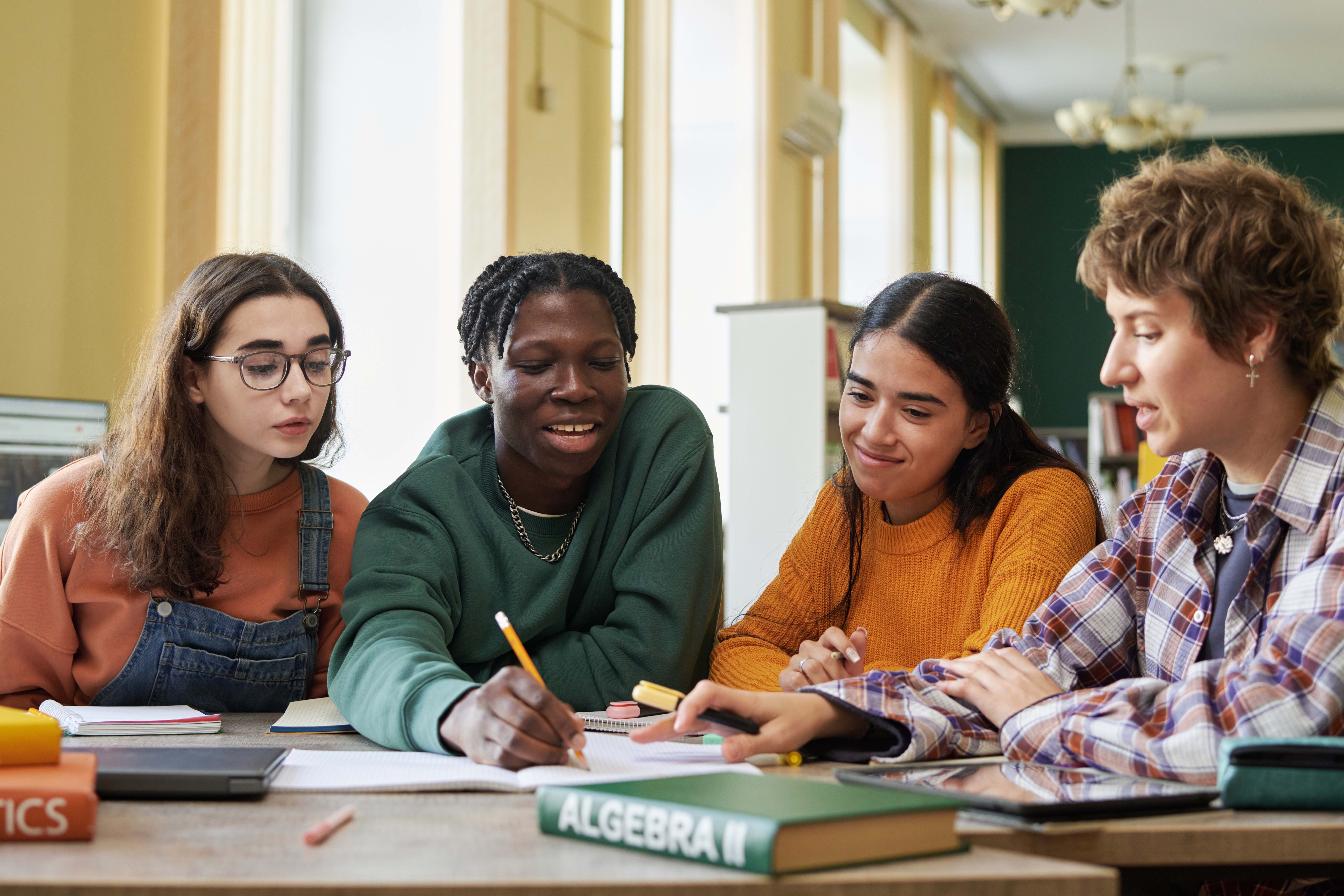 Four diverse high school students studying together at a desk with textbooks, collaborating on schoolwork in a classroom