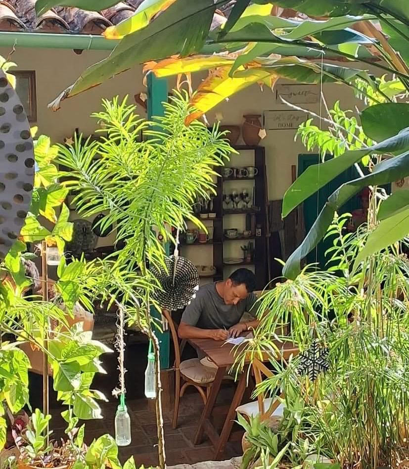 Hombre sentado escribiendo en una libreta en el jardín tropical del café, rodeado de plantas y con una estantería de cerámica al fondo