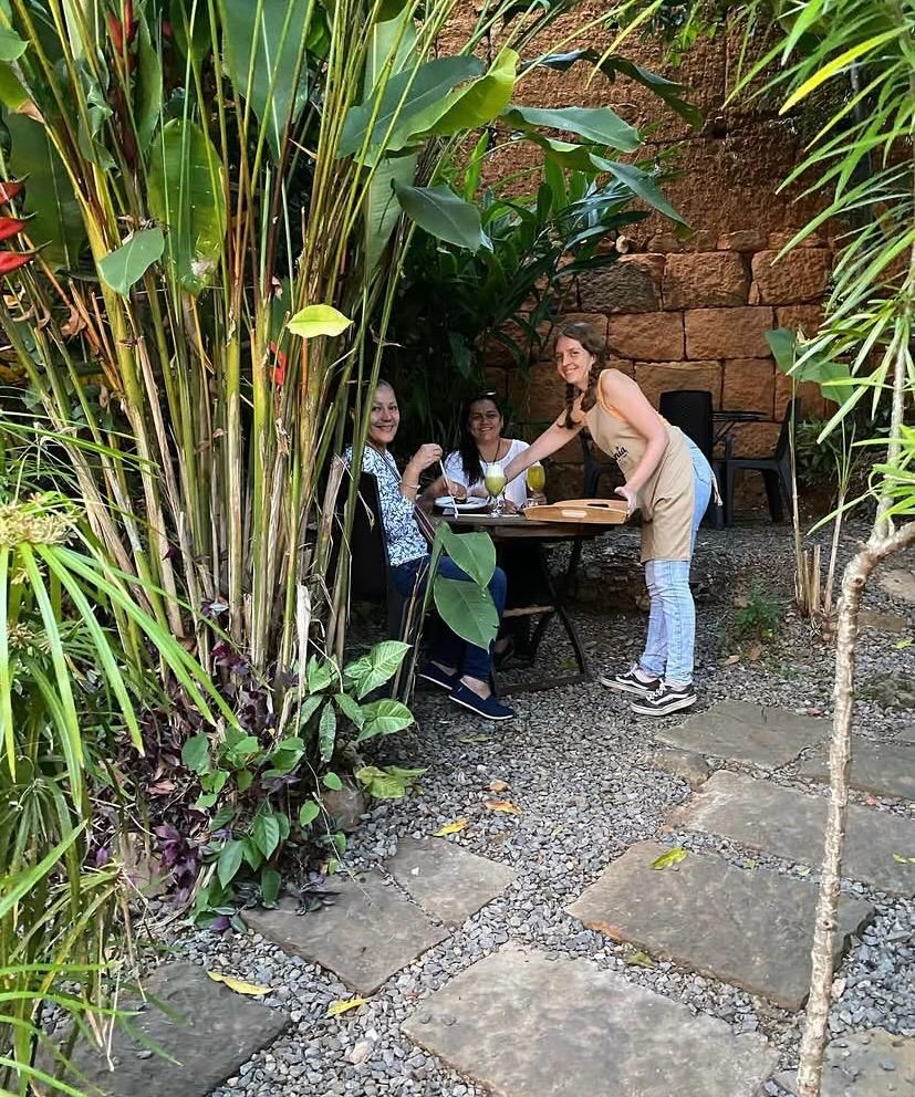 Mesera sirve bebidas a dos clientes sentadas en el frondoso jardín tropical de El Ángel Café, con un muro de piedra arenisca al fondo