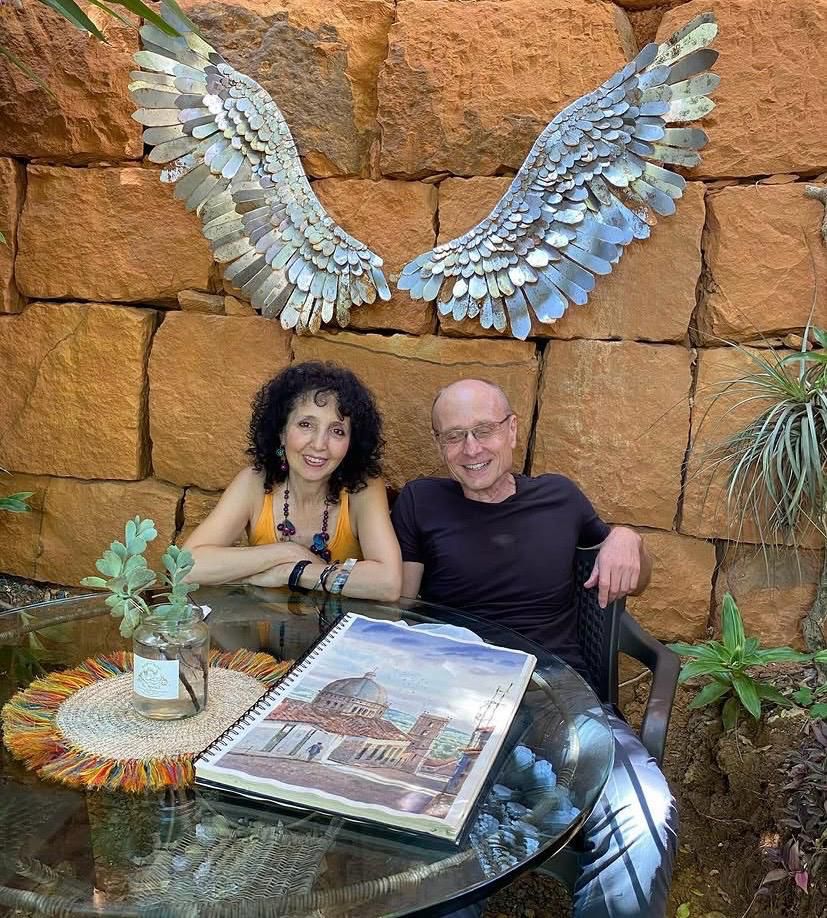 Pareja sonriendo en el jardín del café, con una escultura de grandes alas metálicas detrás y un libro de acuarelas de Barichara sobre la mesa
