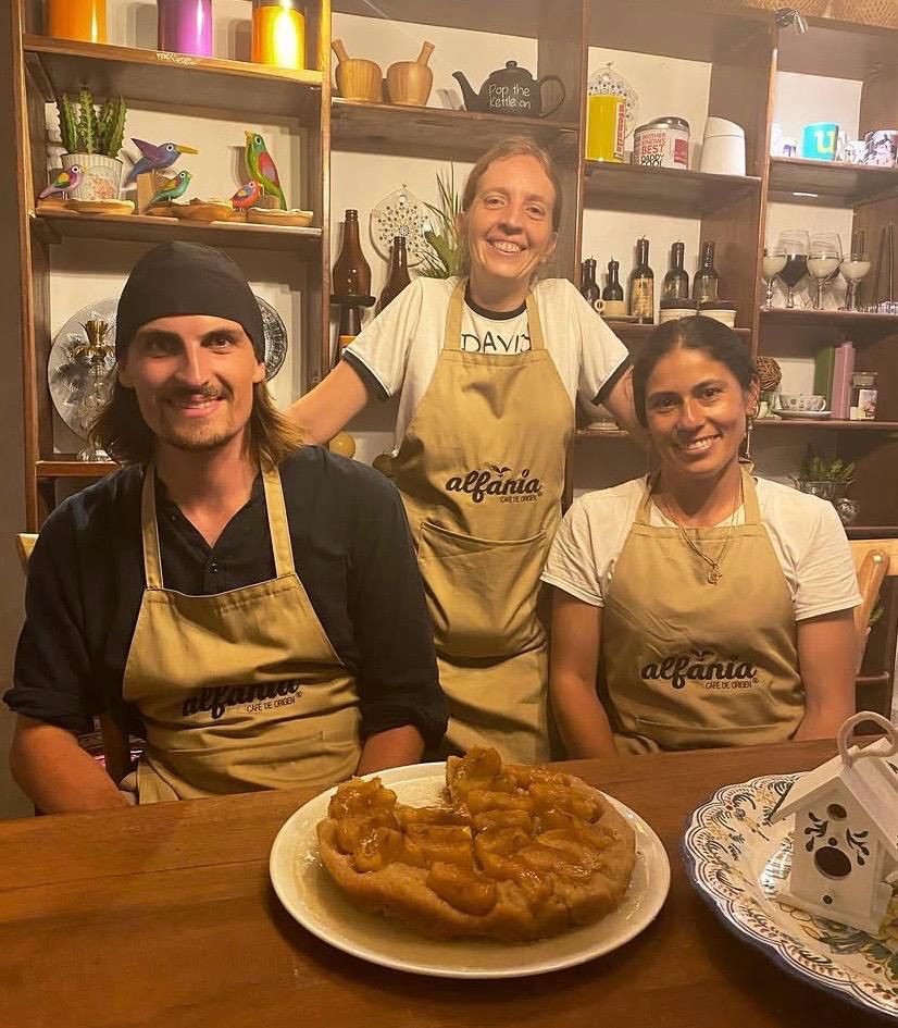 Tres personas con delantales de Alfanía Café de Origen sonriendo detrás de una mesa con una tarta de manzana caramelizada recién horneada