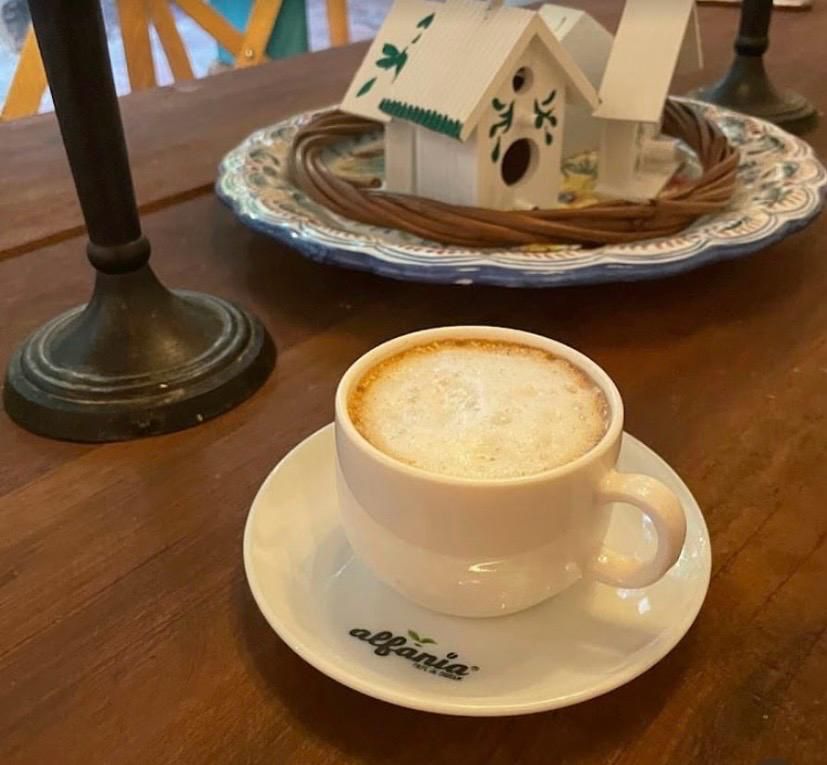 Taza de café Alfanía con leche espumada sobre plato de cerámica artesanal, con una decoración de casitas de pajaritos al fondo