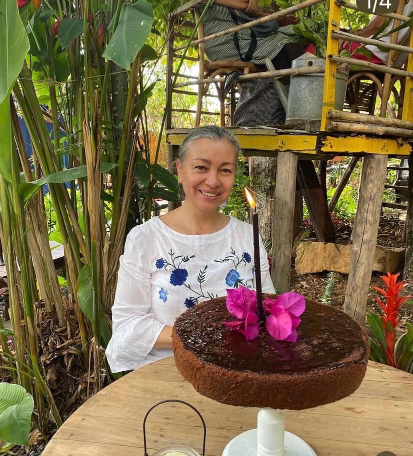 Mujer sonriendo con una torta de chocolate decorada con flores de bugambilia y una vela encendida, en el jardín del café entre heliconias y bambú
