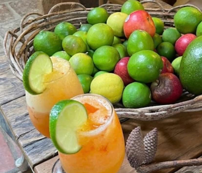 Dos vasos con jugo de mandarina y tajada de limón sobre una mesa rústica, junto a una canasta llena de frutas tropicales frescas