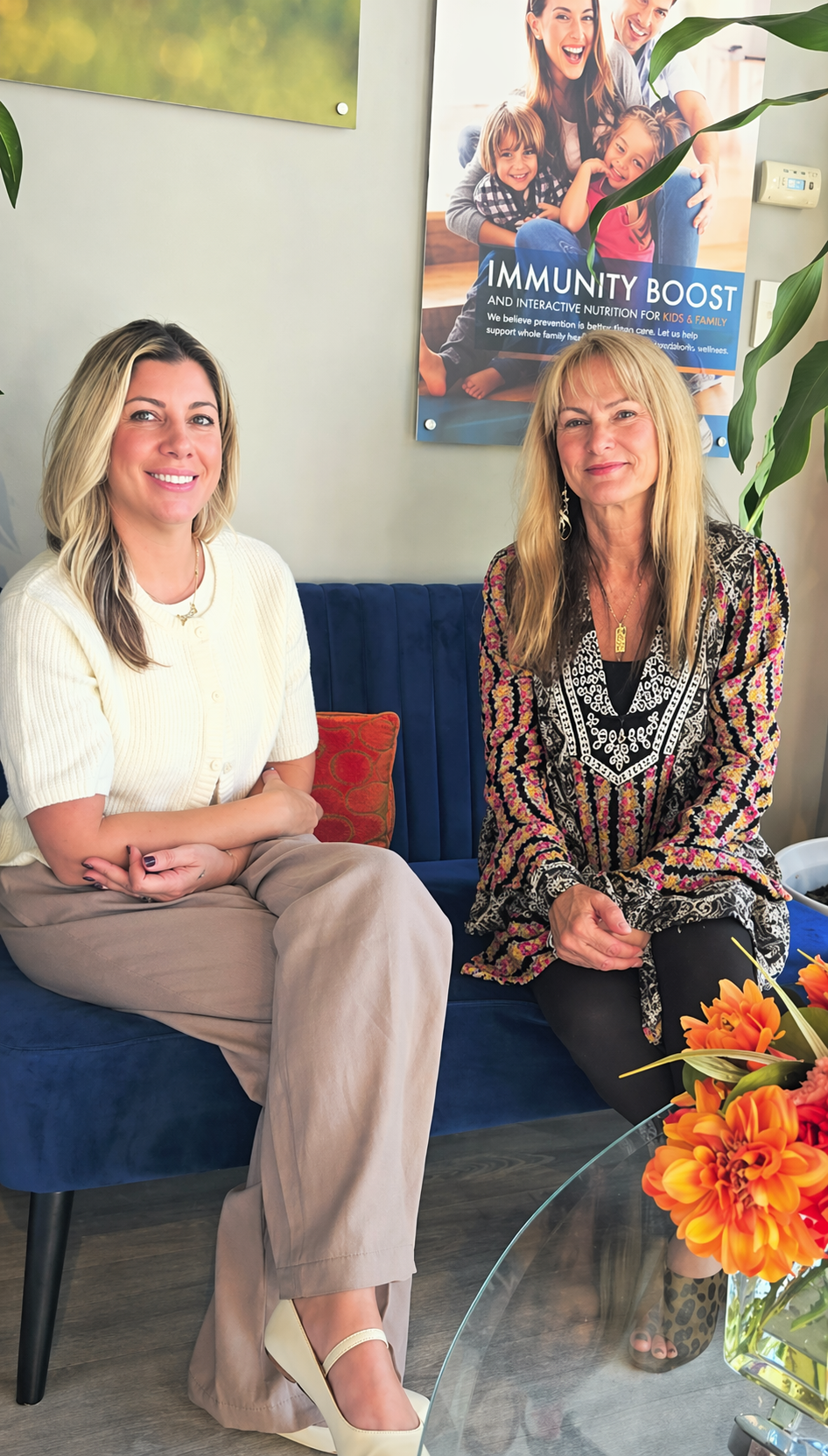 Two women smiling on a navy couch beneath an Immunity Boost poster